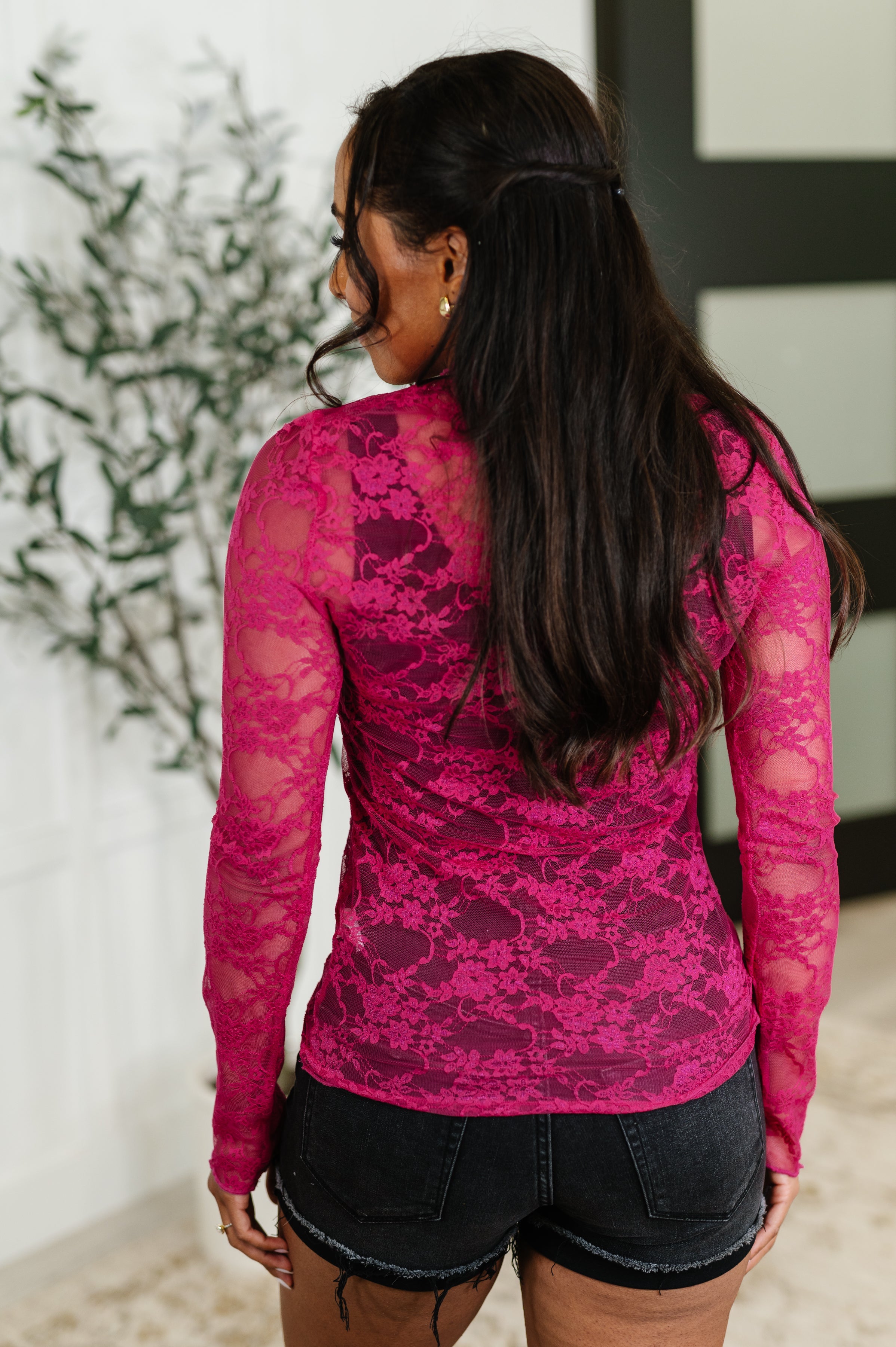 Woman with long dark hair wearing fitted fuchsia lace layering top and black denim shorts indoors near plant