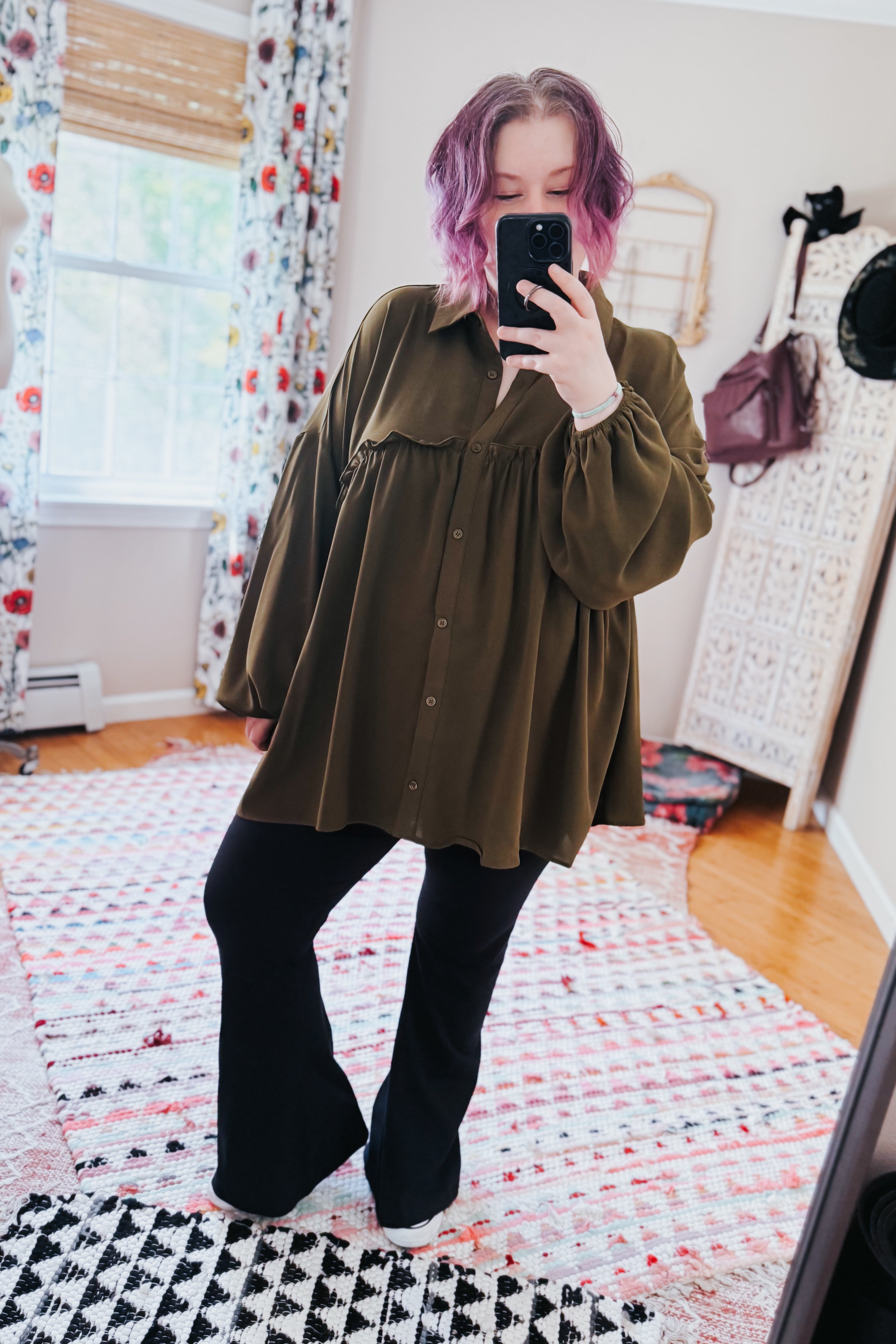 Woman with purple hair wearing loose olive button-down blouse paired with black flared pants taking mirror selfie indoors