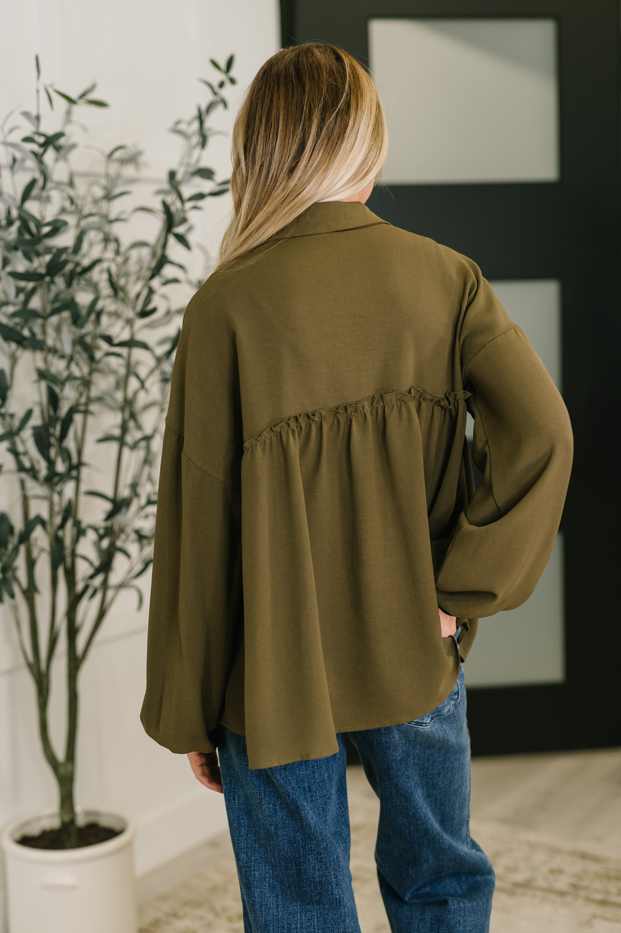 Back view of olive button down blouse with gathered detail, worn with blue jeans in indoor setting with plant nearby