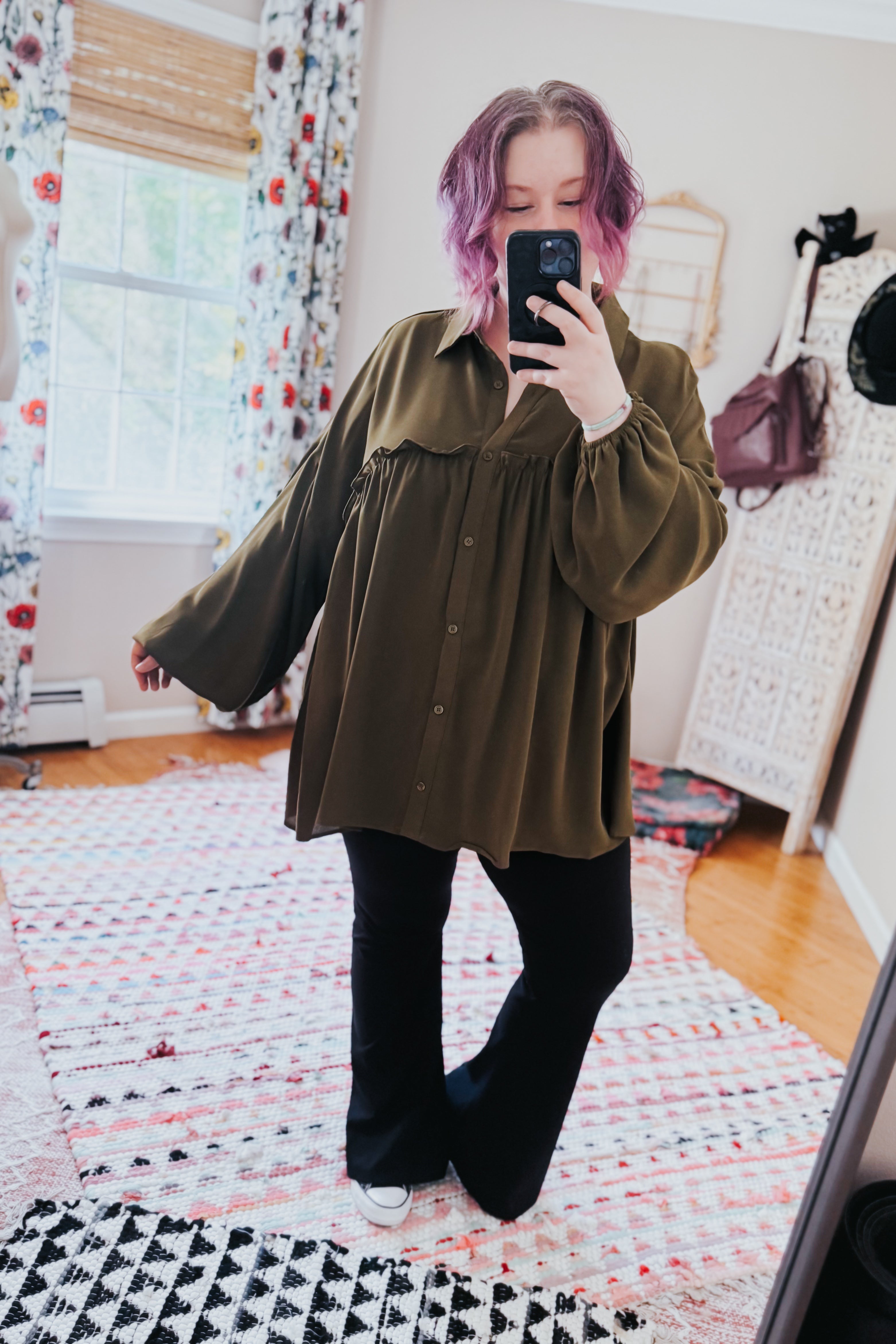 Woman with purple hair wearing oversized olive button-down blouse paired with black pants and white shoes taking mirror selfie