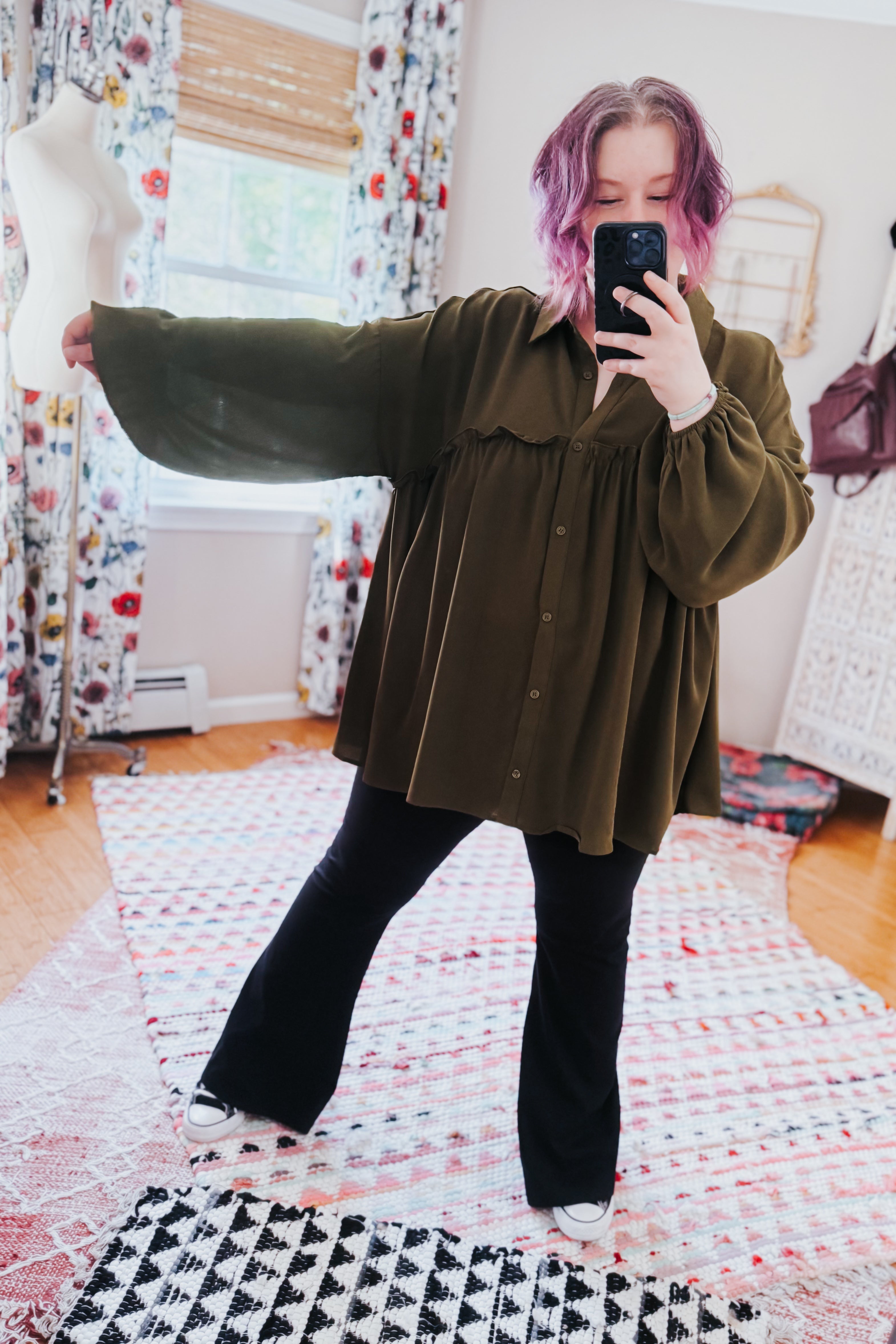 Woman with purple hair wearing oversized olive button-down blouse and black pants taking mirror selfie indoors