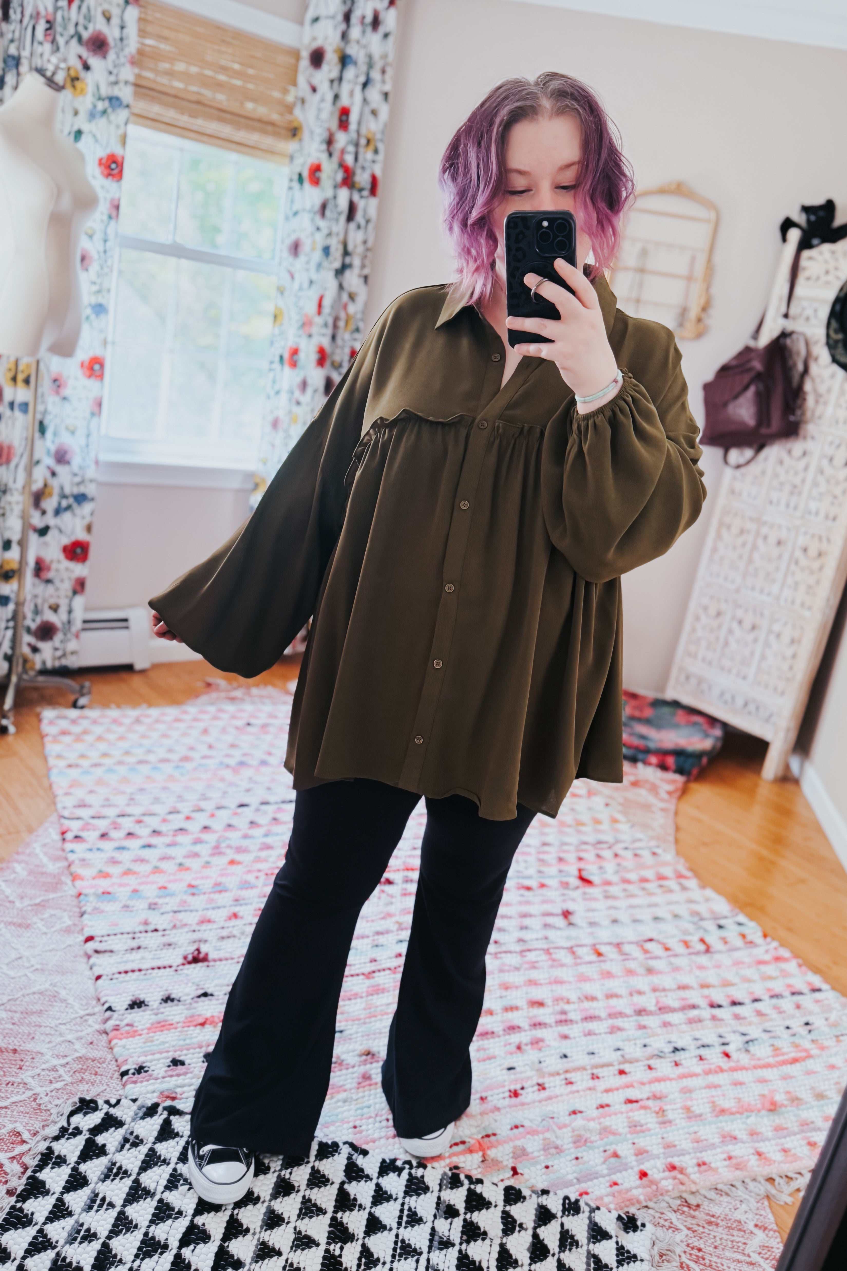 Woman with purple hair wearing loose olive button-down blouse paired with flared black pants taking mirror selfie indoors
