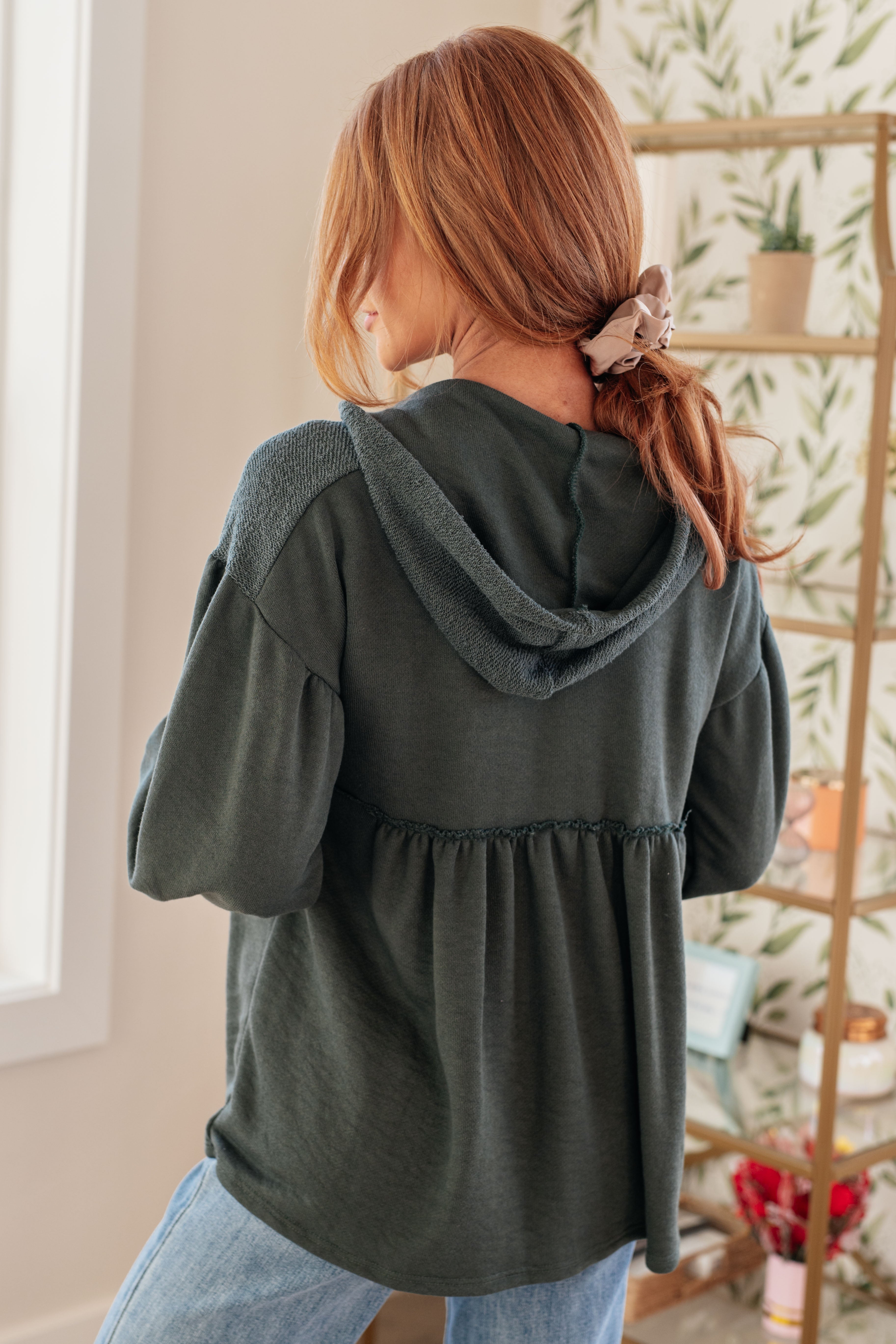 Woman wearing a dusty green v-neck hooded pullover and blue jeans in an indoor setting.