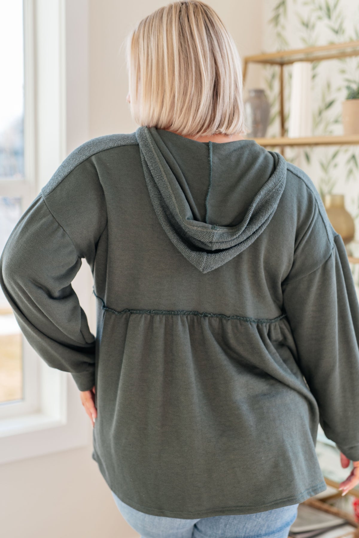 Woman viewed from behind wearing a teal hooded pullover with a gathered peplum and balloon sleeves over light wash jeans indoors
