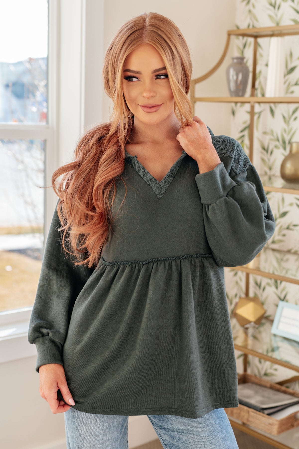 Woman modeling a teal hooded V-neck pullover with gathered peplum and balloon sleeves styled with light wash jeans indoors