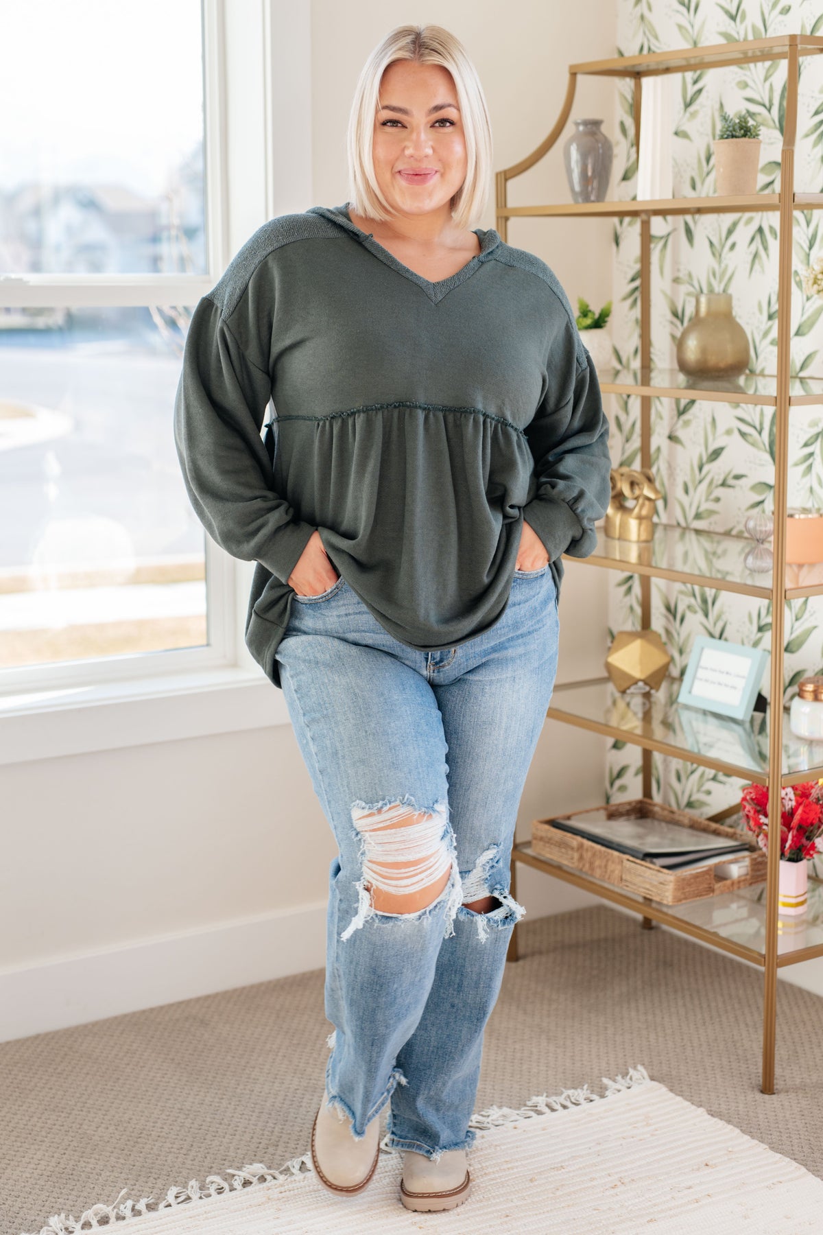 Woman modeling a teal hooded V neck pullover with gathered peplum and balloon sleeves, paired with distressed jeans indoors