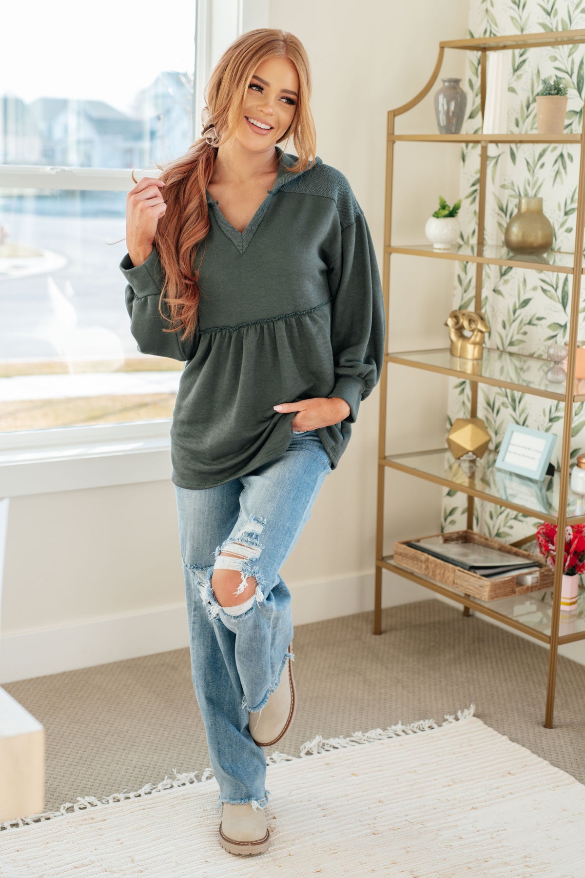 Smiling woman modeling a teal hooded V neck pullover with gathered peplum and balloon sleeves paired with distressed jeans indoors