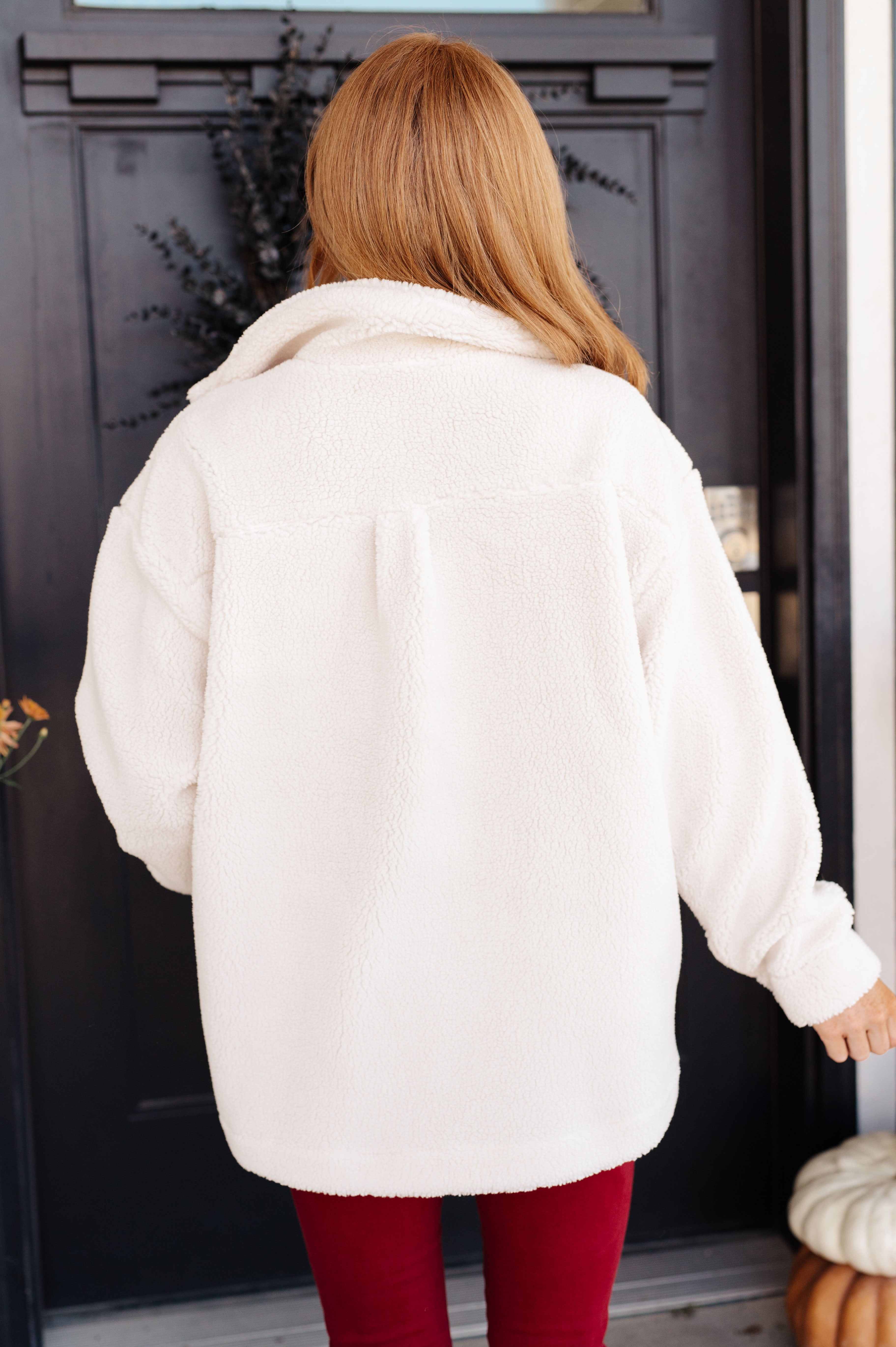 Woman wearing oversized white sherpa coat with relaxed fit viewed from back against dark door background