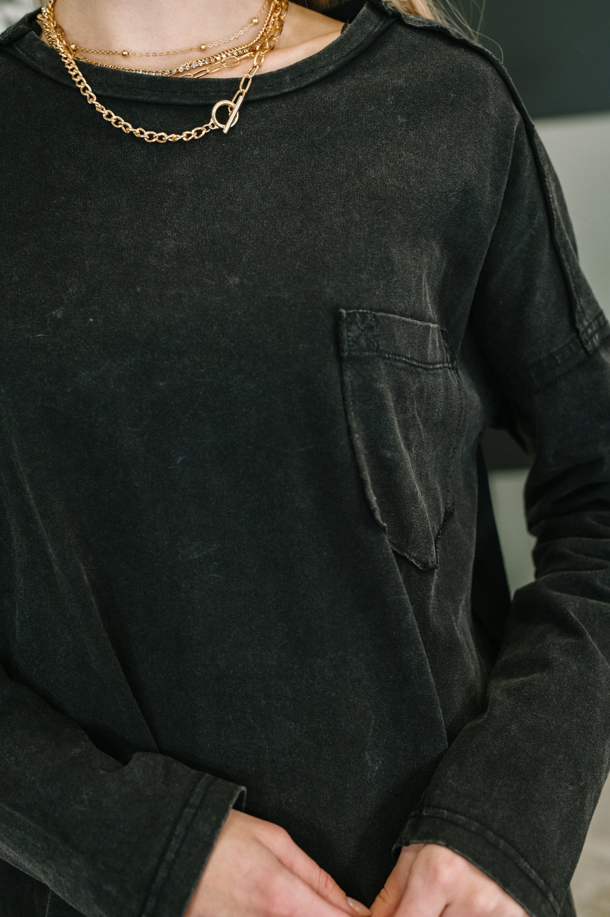 Close view of a person wearing a black long sleeve drop shoulder top with exposed seams chest pocket and layered gold necklaces