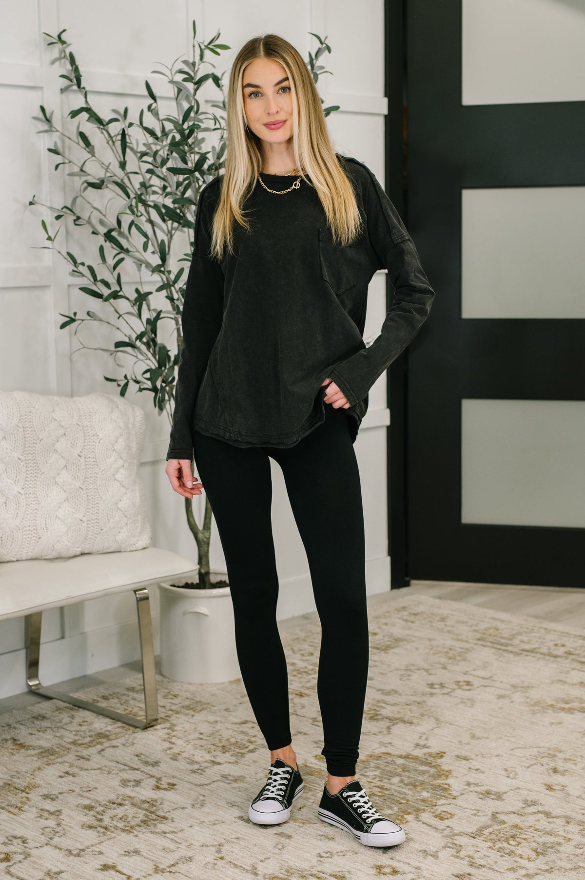 Woman modeling a black drop shoulder top with exposed seams black leggings and casual sneakers in a stylish indoor setting
