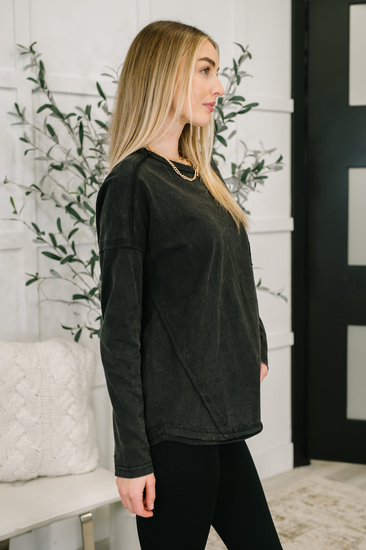 Woman in profile wearing a black long sleeve drop shoulder top with exposed seams and black leggings in a bright home interior