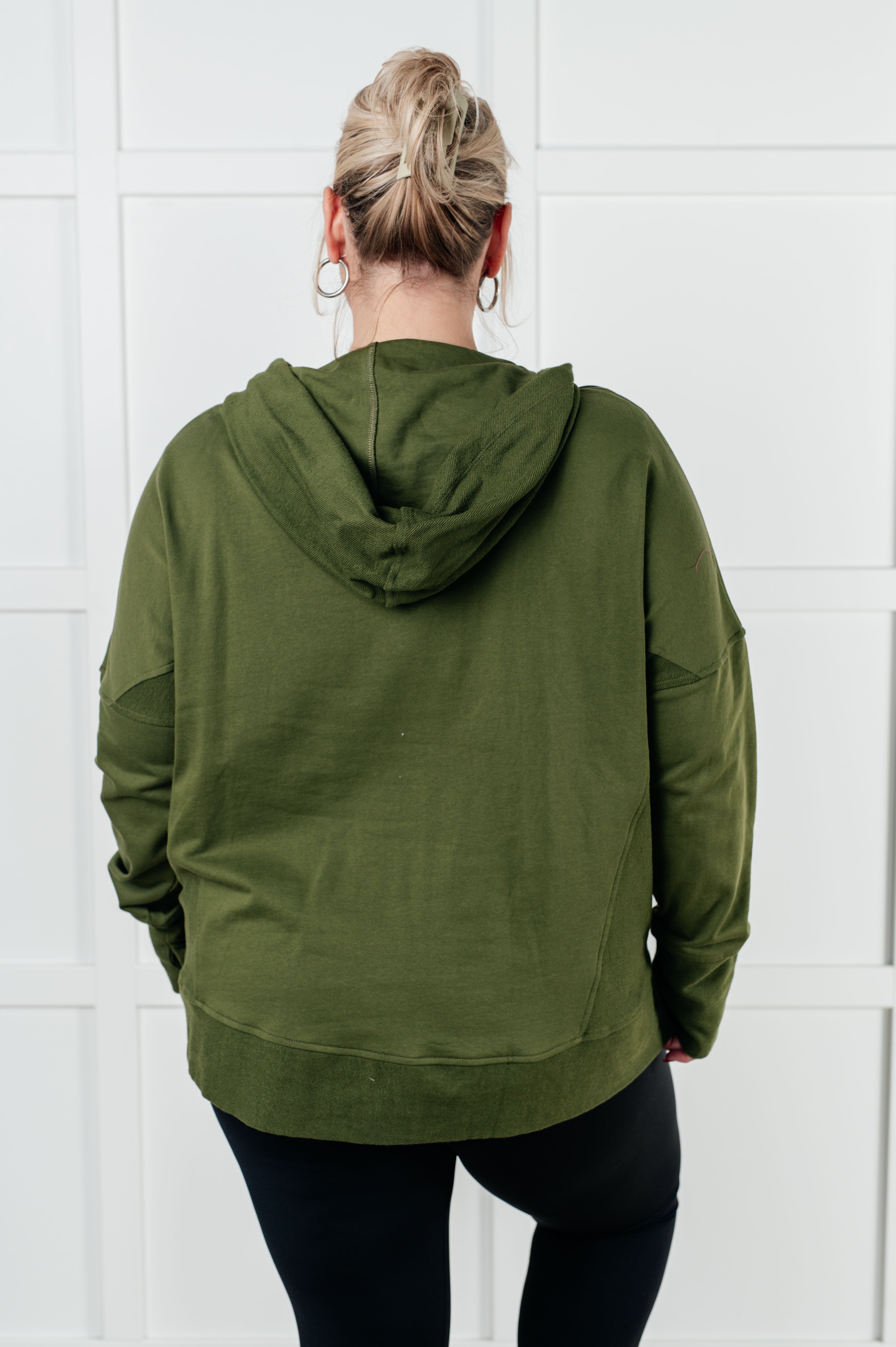 Woman wearing relaxed-fit green hooded pullover viewed from back against white paneled wall