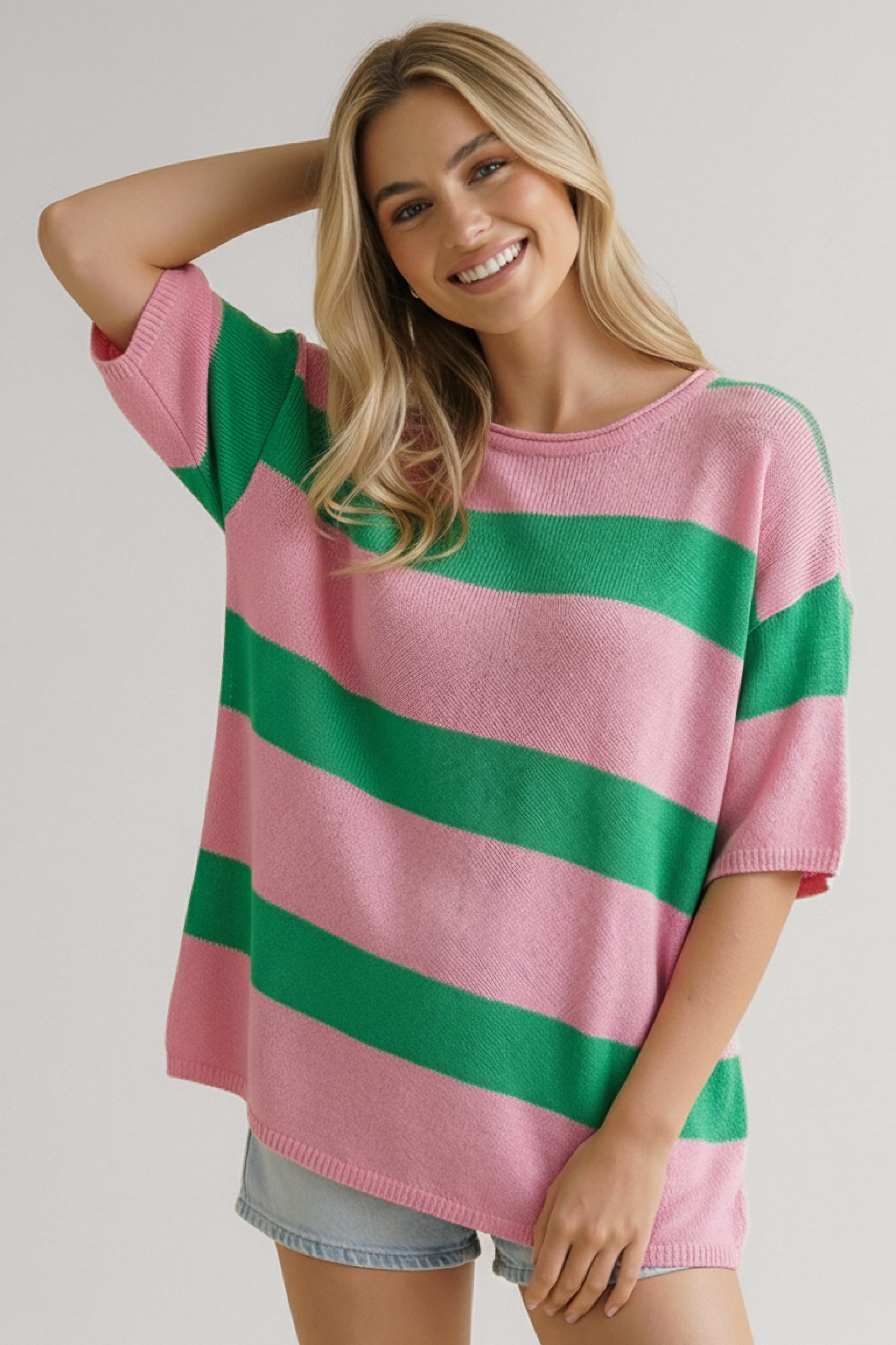 Smiling woman wearing a relaxed pink and green horizontally striped knit top with elbow-length sleeves and light denim shorts