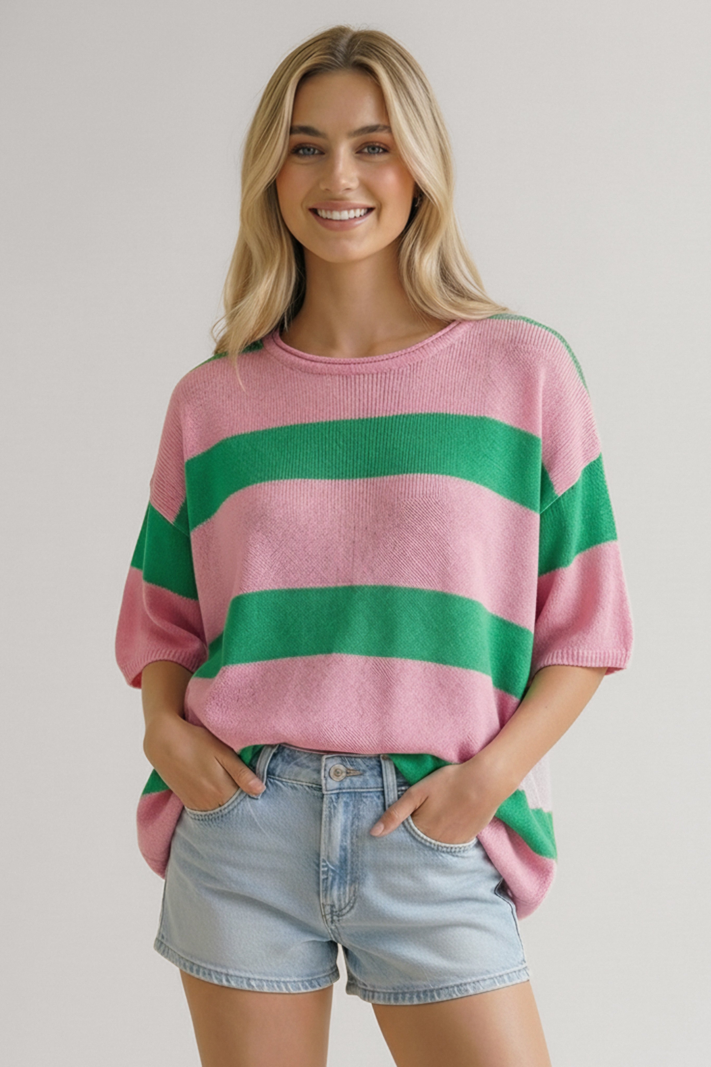 Smiling woman wearing a loose pink and green wide-striped knit top with elbow-length sleeves and light denim shorts