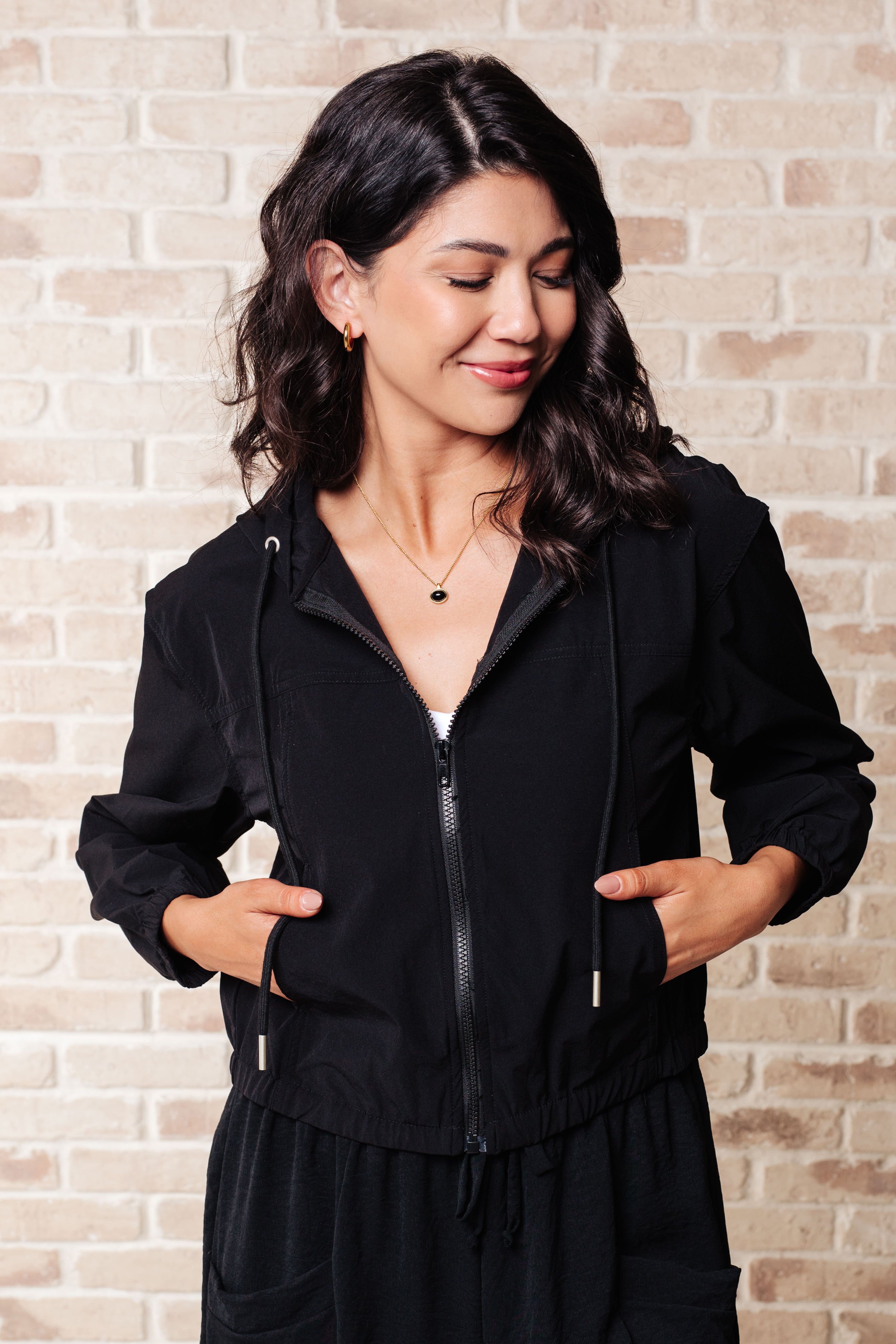 Woman wearing black zip-up hoodie with drawstrings and relaxed fit, smiling with eyes closed against brick wall background
