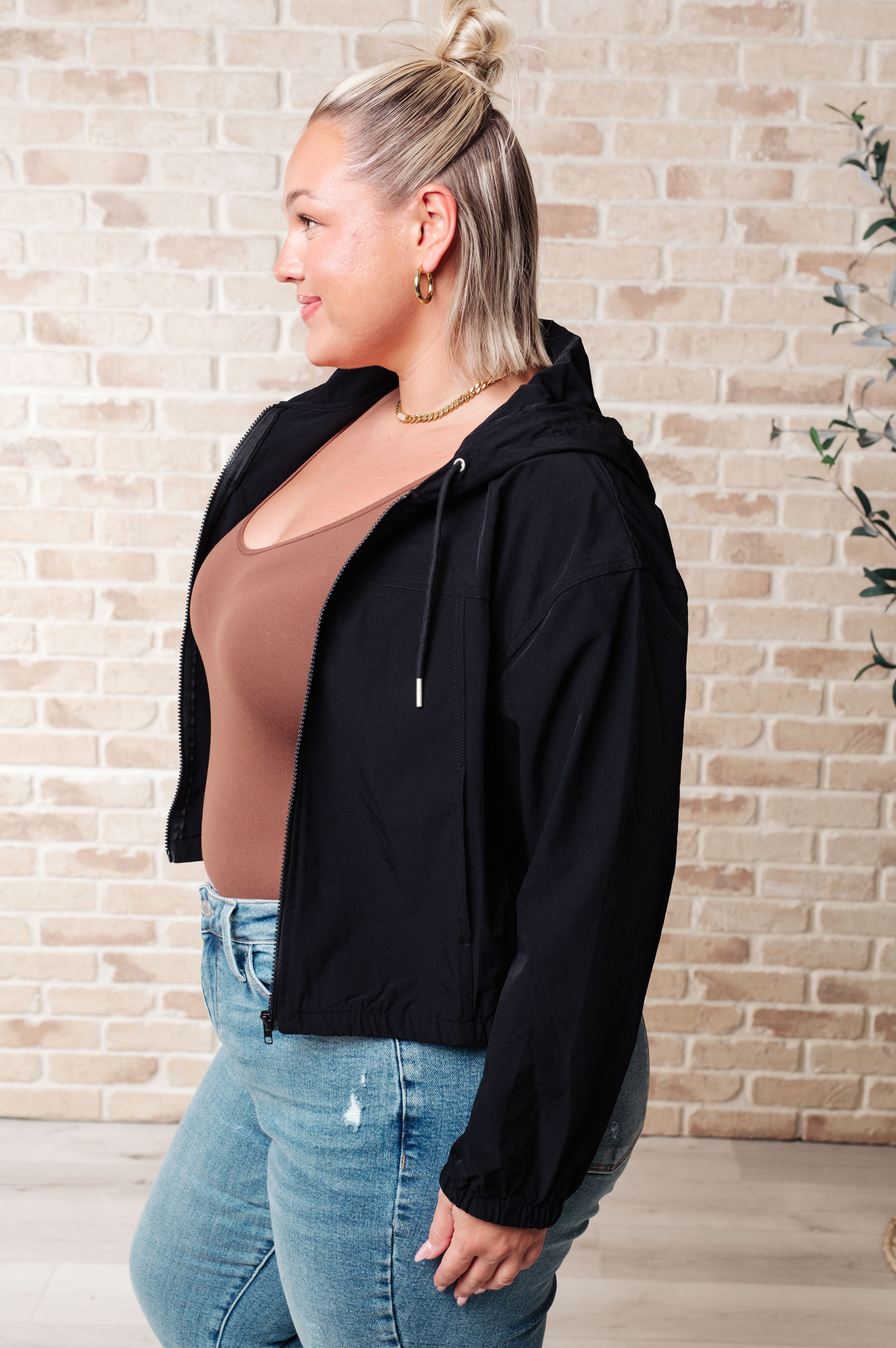Woman wearing black zip-up hoodie over brown top paired with light wash jeans against brick wall background