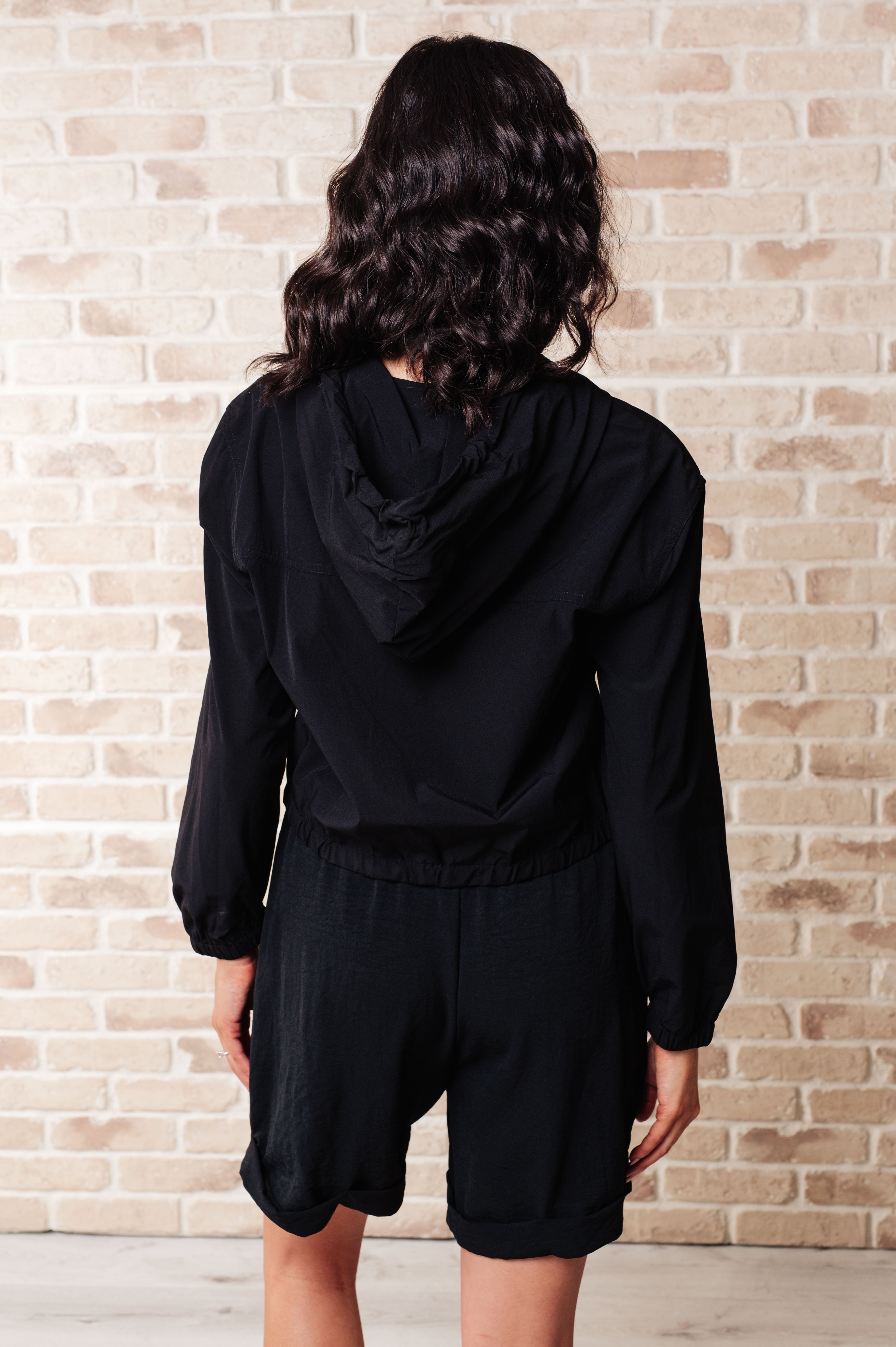 Woman shown from behind wearing black zip-up hoodie with hood and loose black shorts against brick wall