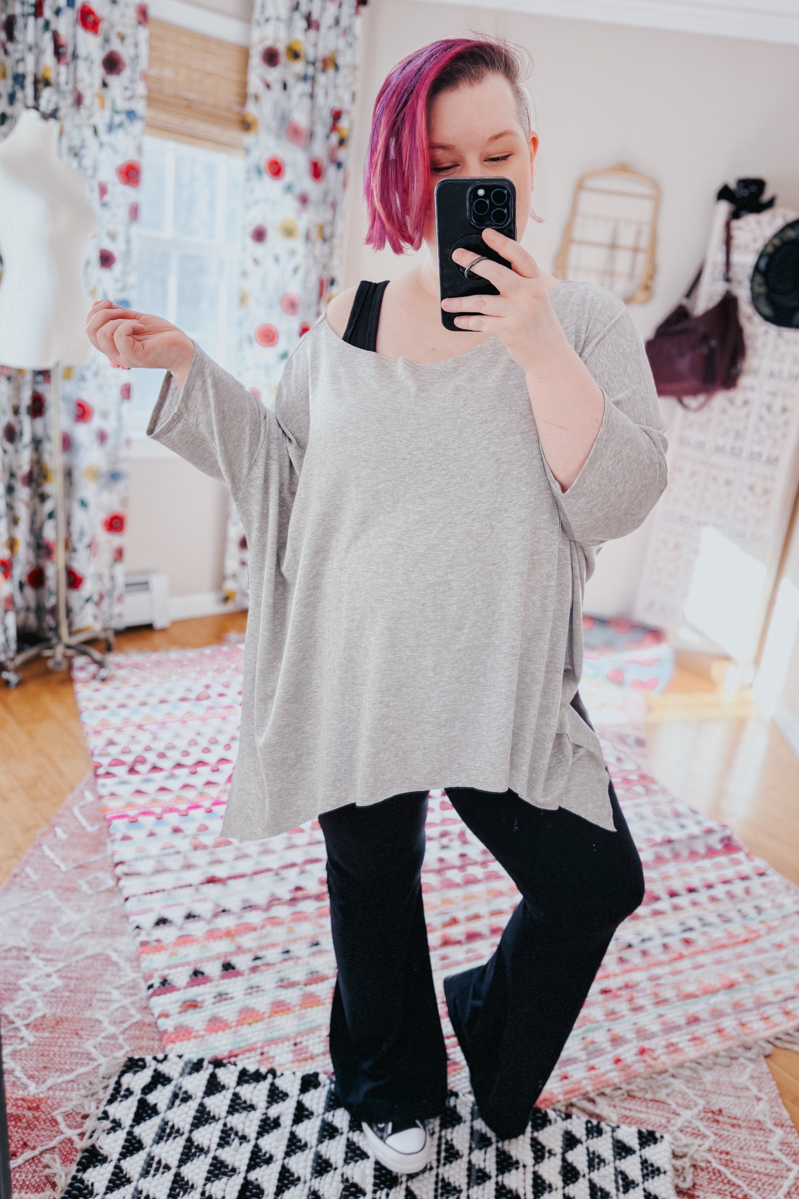 Woman with pink hair wearing loose grey flowy top and black pants taking mirror selfie in bright room with patterned rugs