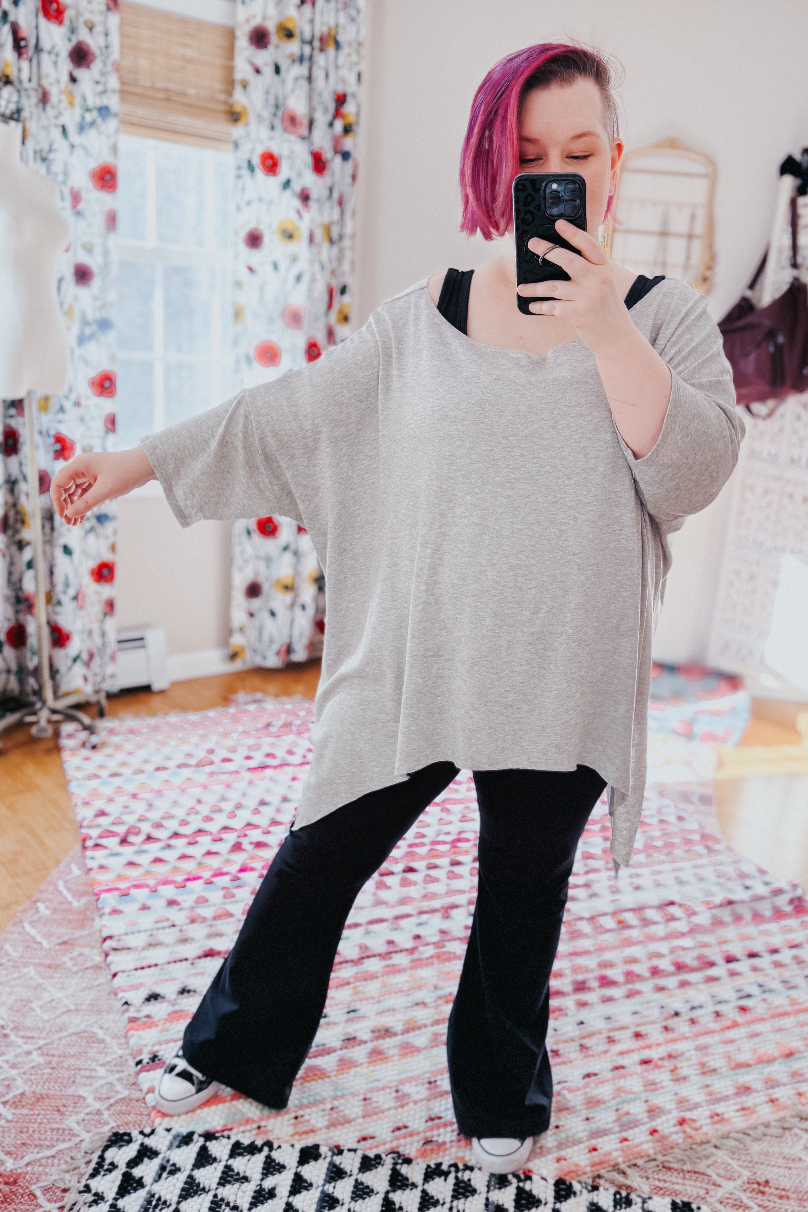 Woman with pink hair wearing loose grey flowy top with raw edge and black pants taking mirror selfie indoors