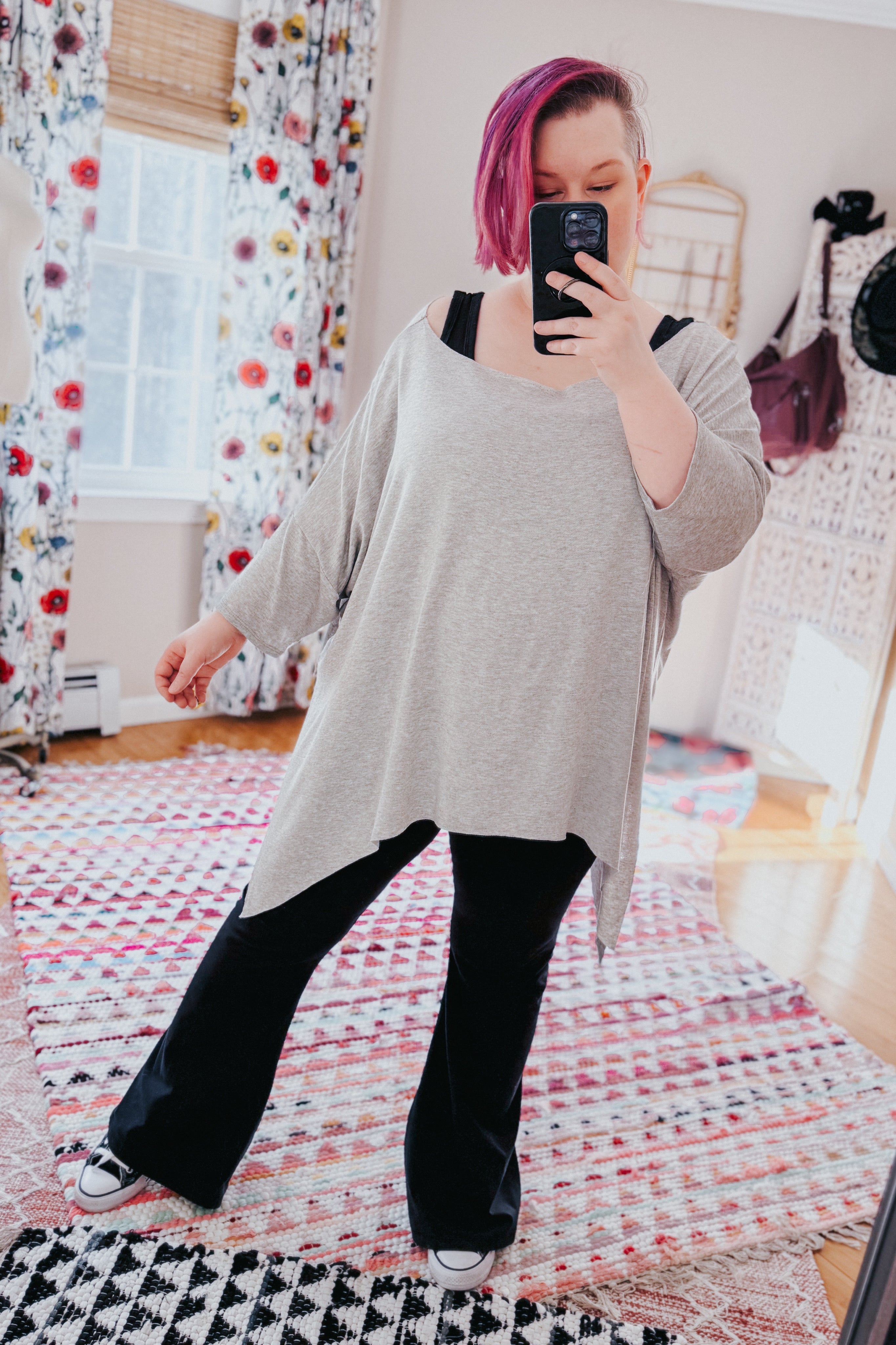 Woman with purple hair wearing a grey flowy top with raw edge hem and black pants taking mirror selfie indoors