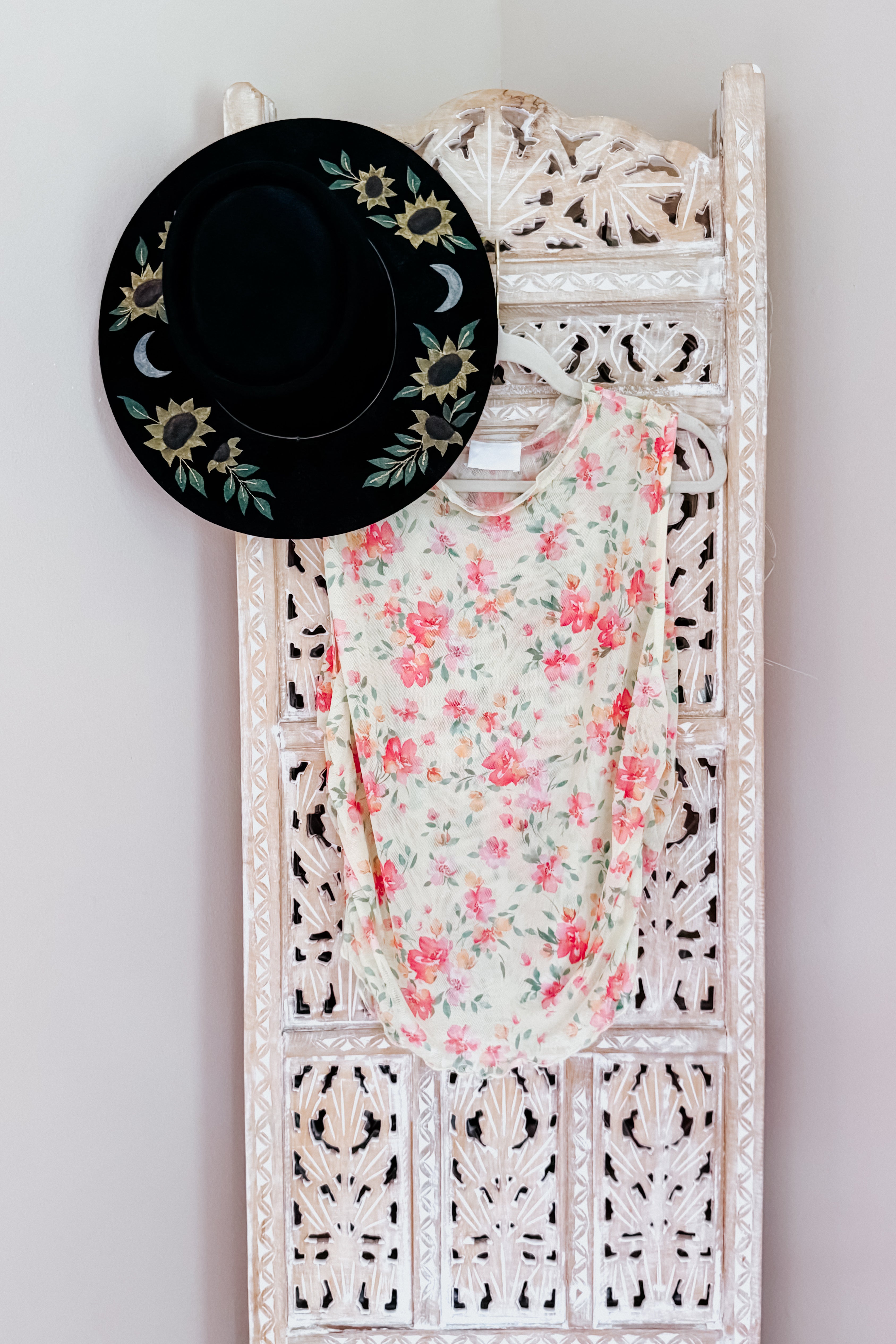 Sleeveless sheer floral top with pink and green pattern hanging on carved white wooden screen with black floral hat