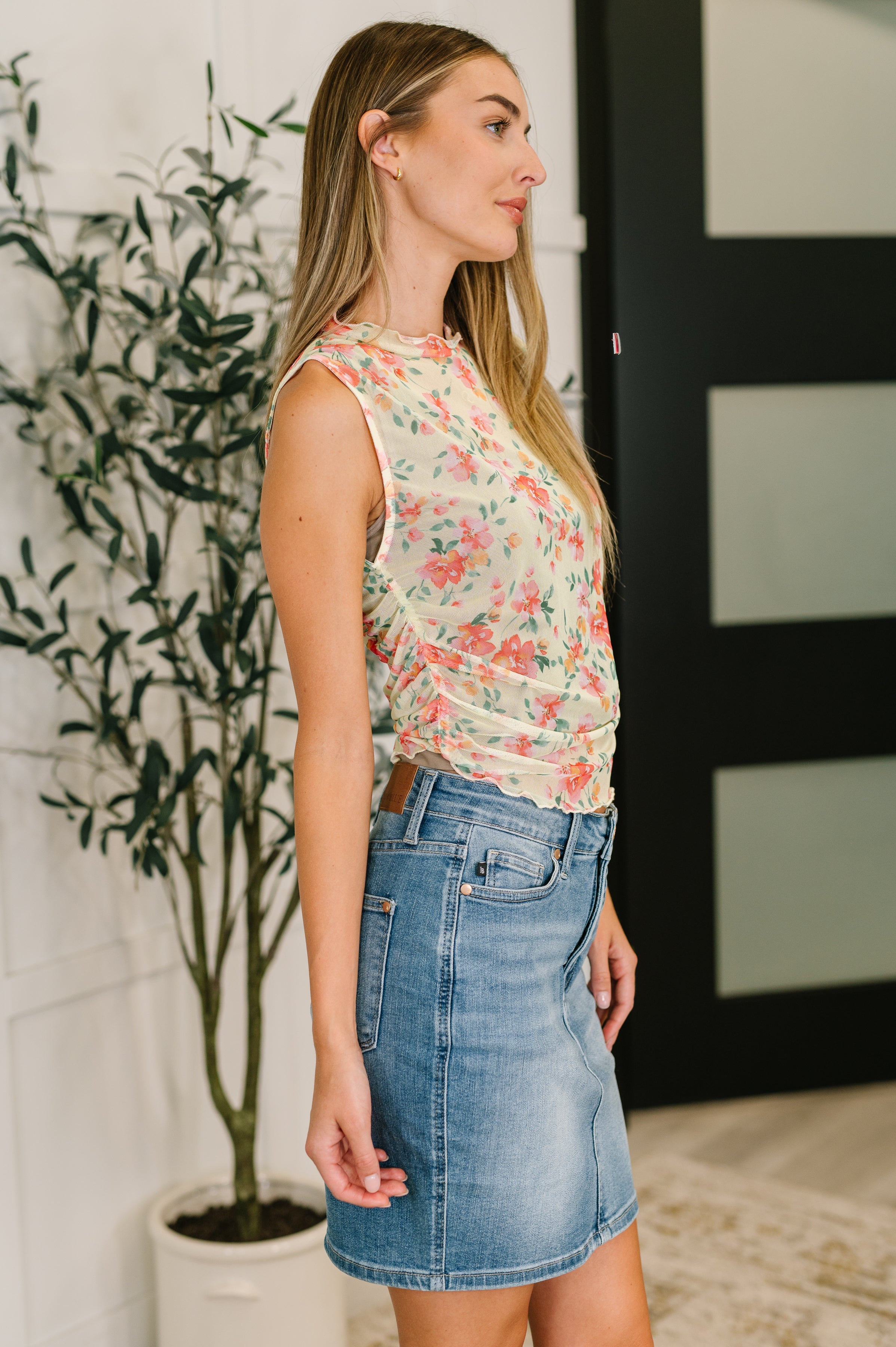Woman wearing sleeveless floral sheer layering top in chartreuse paired with denim skirt indoors near potted plant