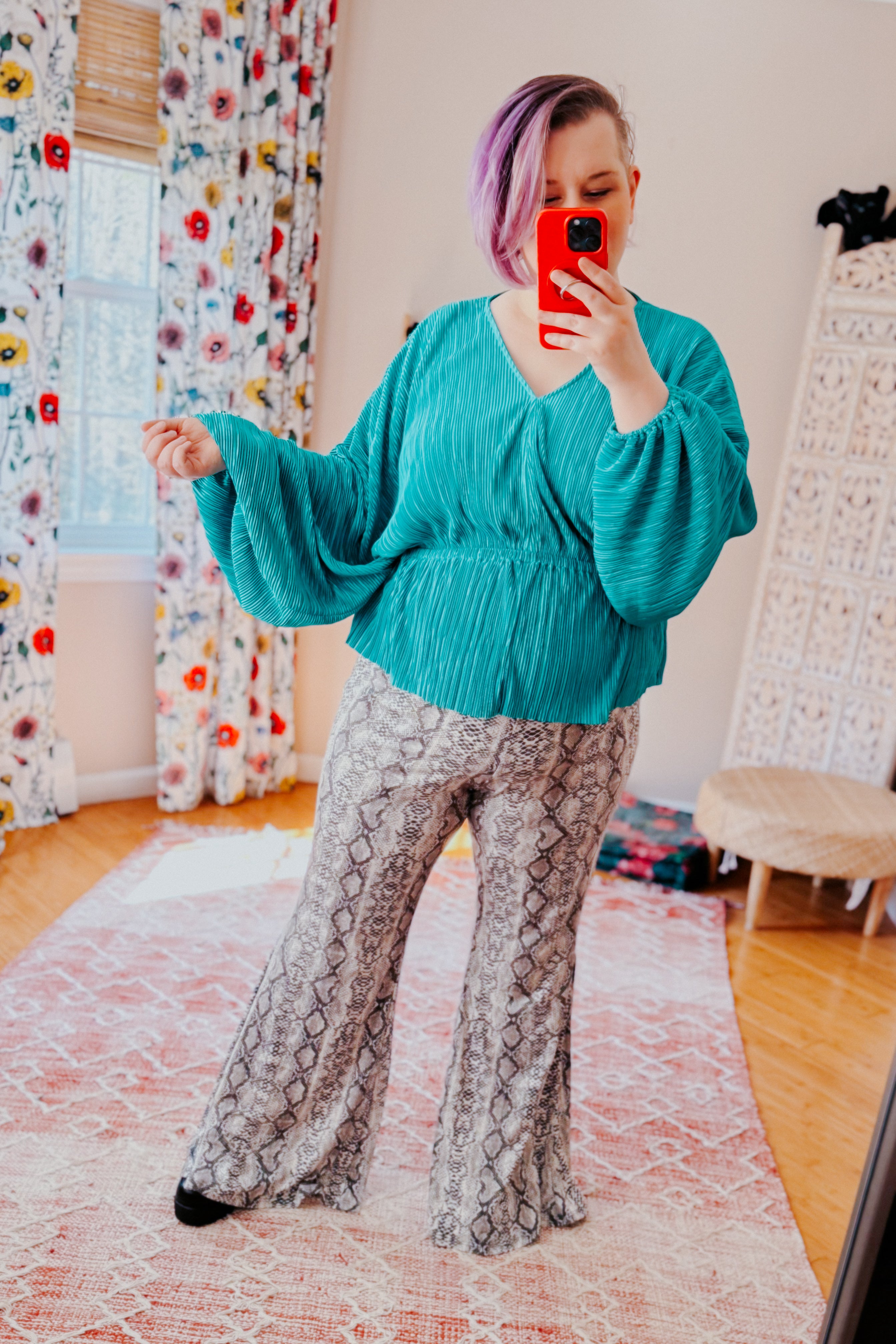Woman with purple hair wearing teal pleated blouse and grey flare pants with snake print taking mirror selfie indoors