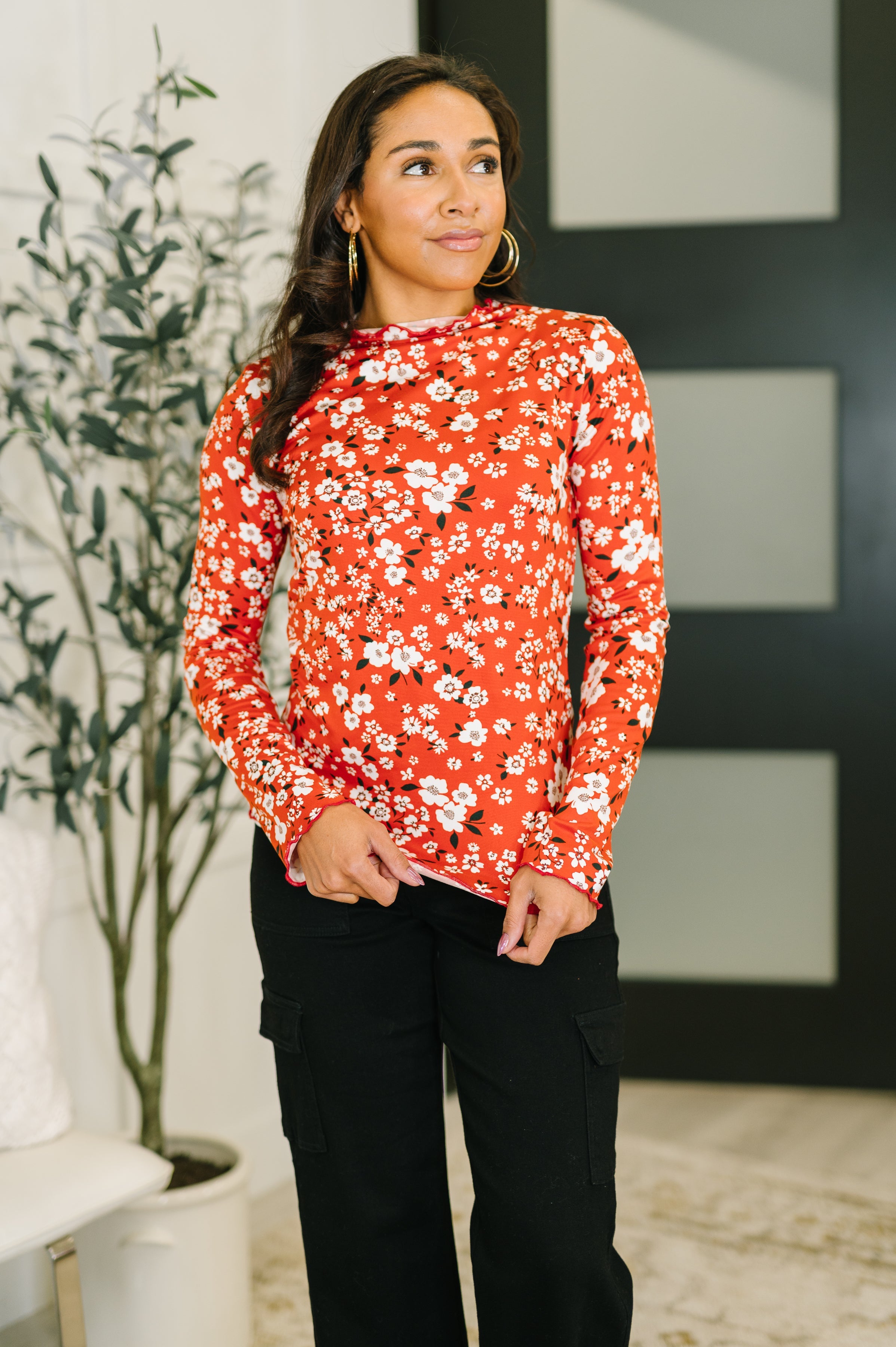 Woman wearing a vermillion floral long sleeve top with a lettuce hem and black pants while posing indoors