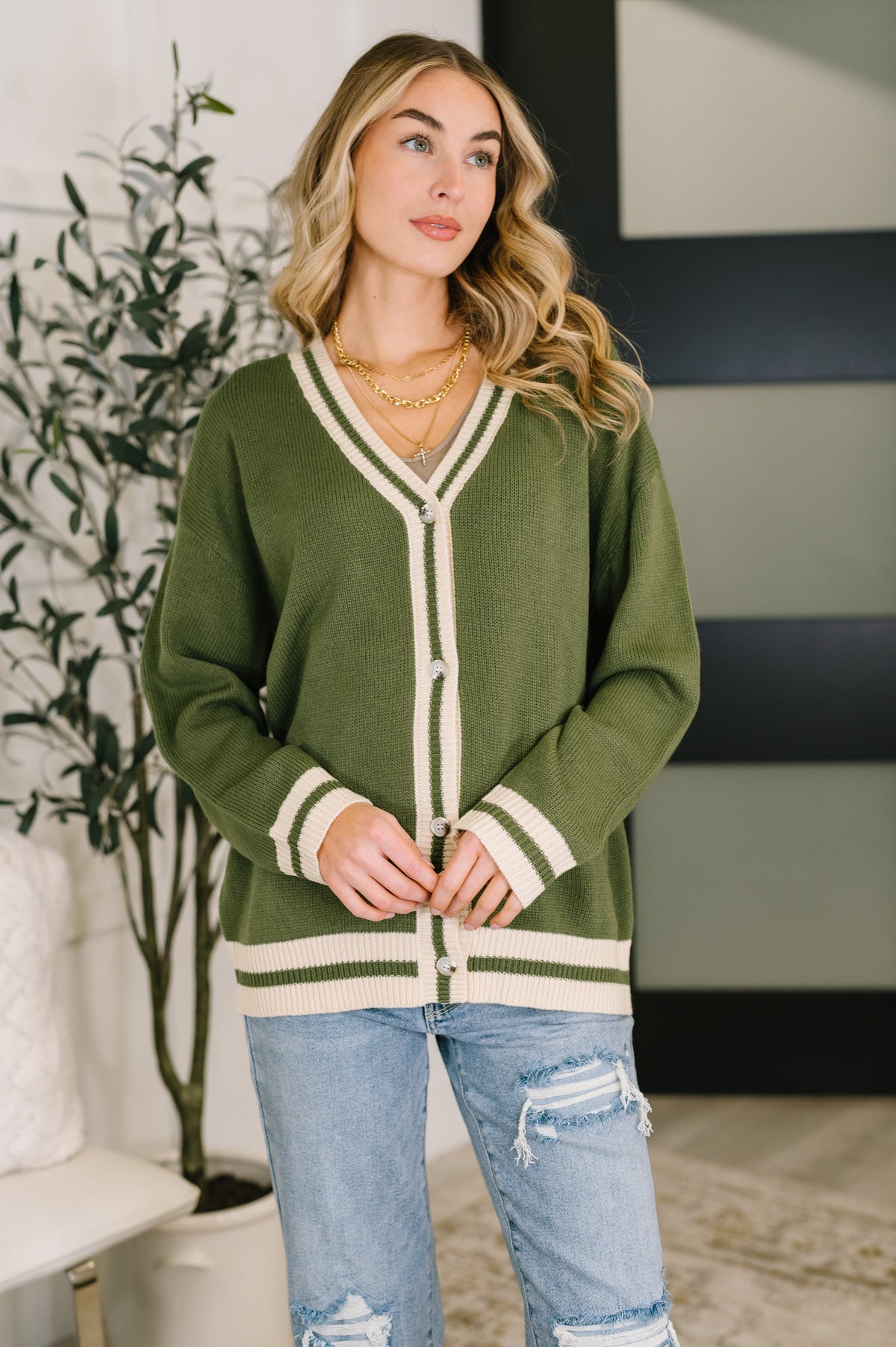 Woman wearing a green button front cardigan with striped ribbed trim, layered necklaces, and distressed jeans in a modern home interior