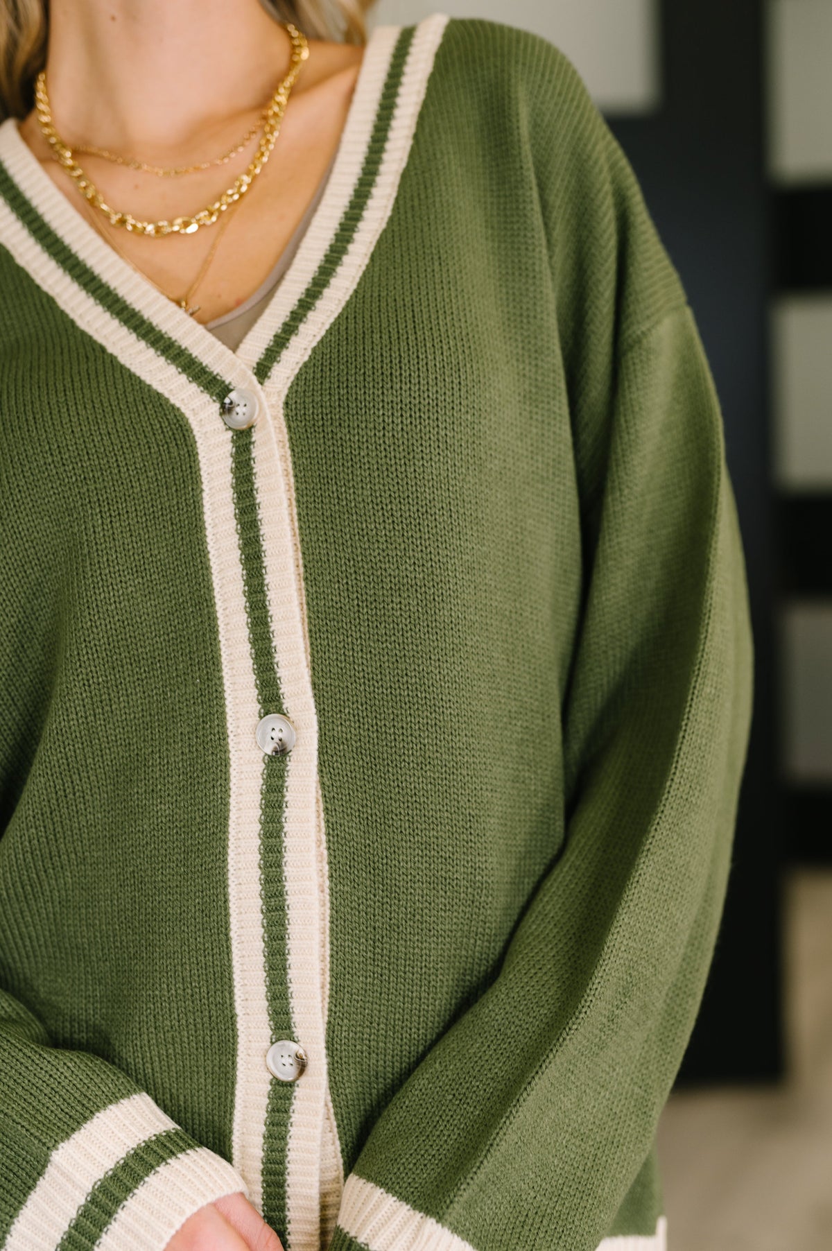 Close view of a woman wearing a green button front cardigan with cream striped trim and layered gold necklaces
