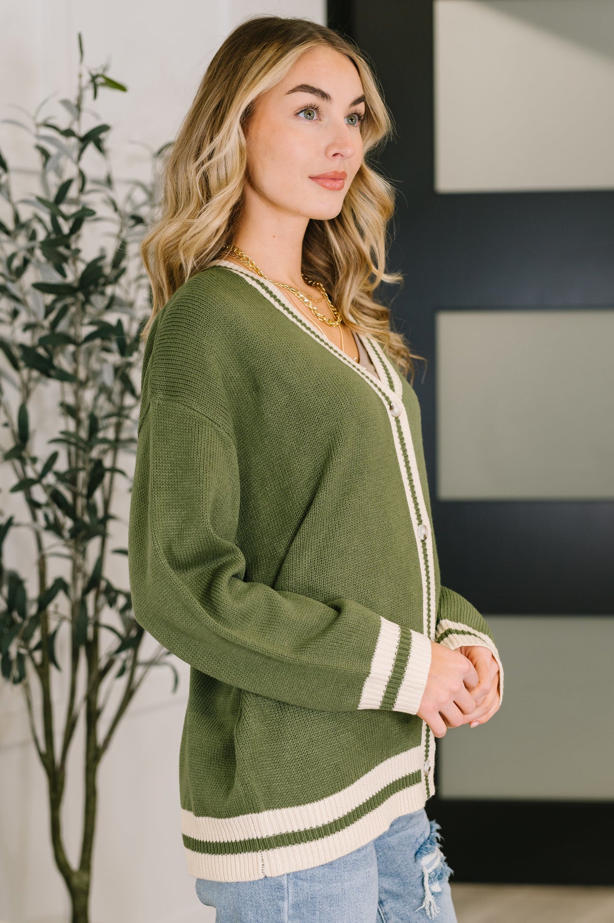 Woman wearing a green button front cardigan with cream striped trim and light blue jeans in a modern home interior