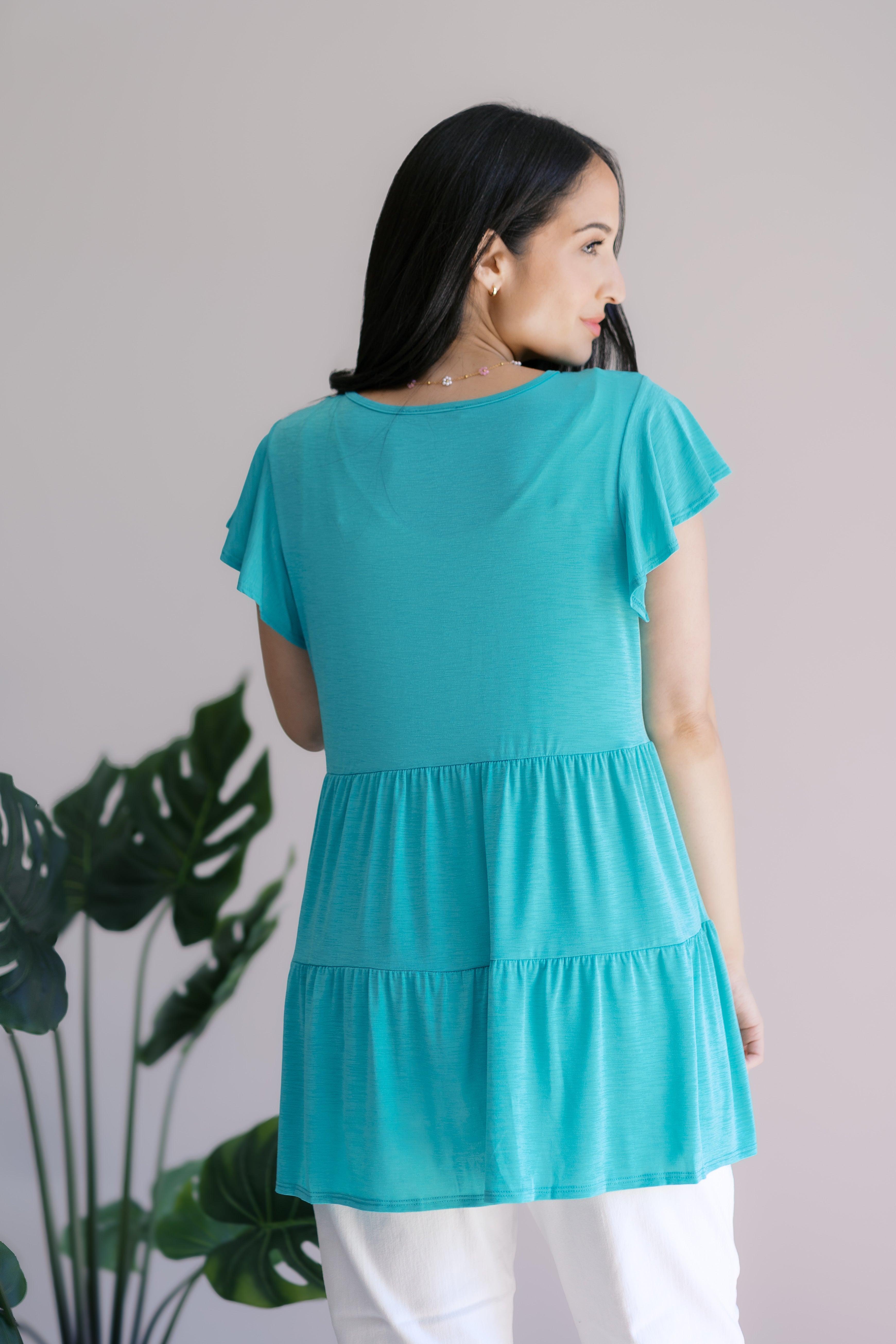 Woman seen from behind wearing a teal babydoll top with flutter sleeves standing indoors near plants