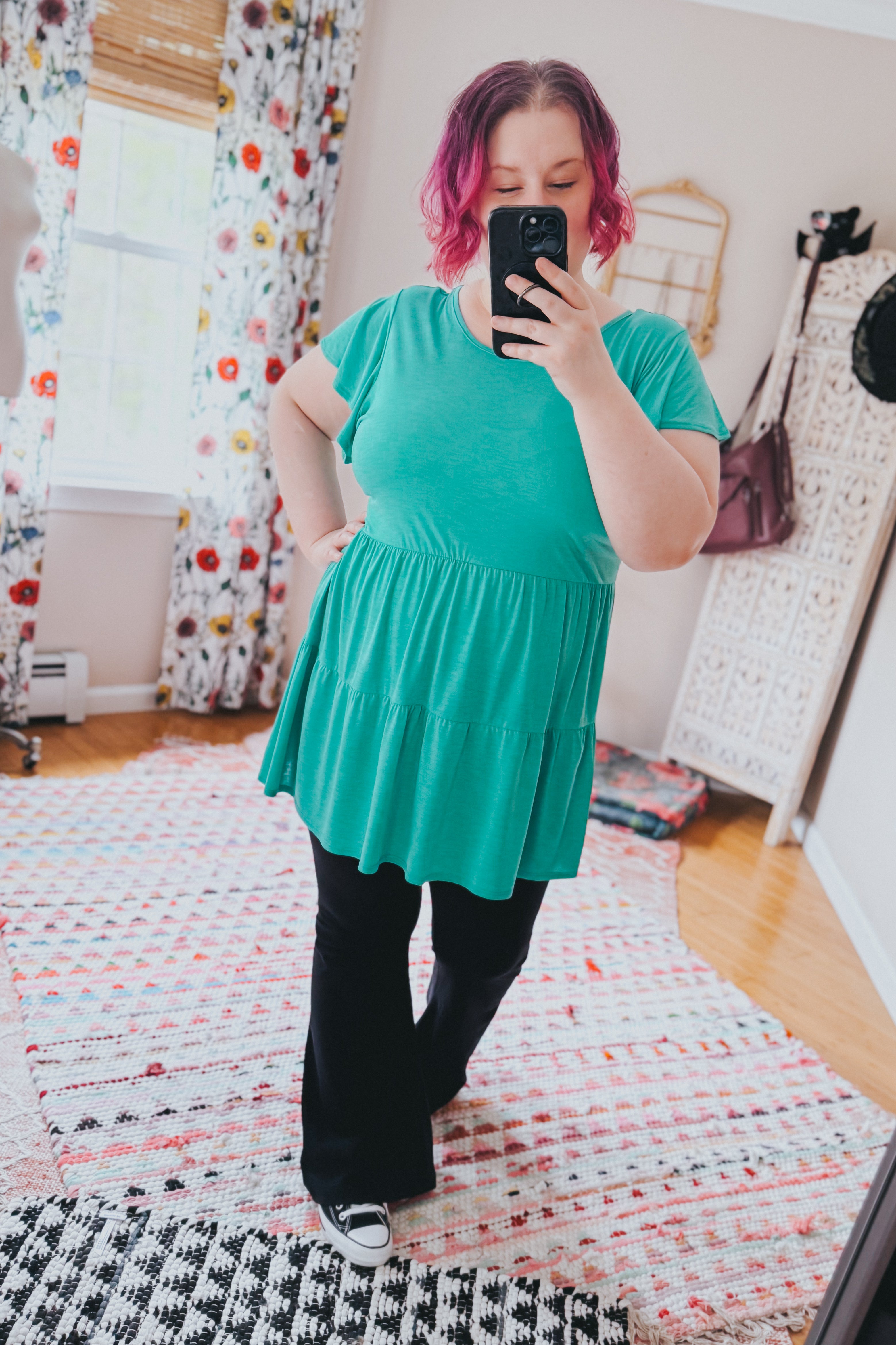 Woman wearing teal babydoll top paired with black pants and sneakers taking mirror selfie indoors