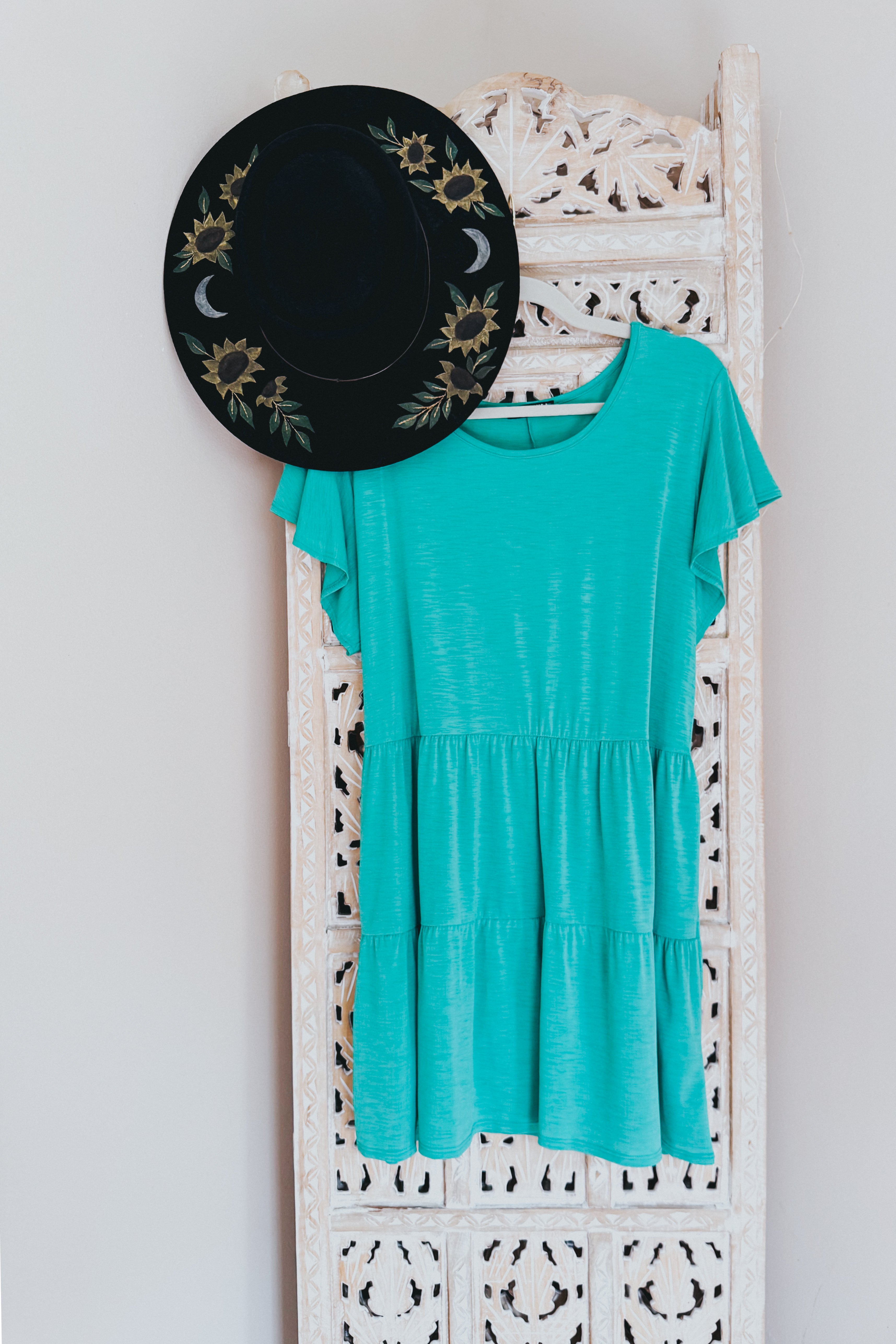 Teal babydoll top with flutter sleeves hanging on carved white wooden panel next to black hat with floral and moon designs