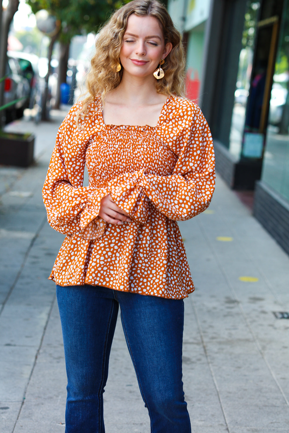 Woman wearing rust smocked top with ruffle hem and puffed sleeves standing outdoors on city sidewalk