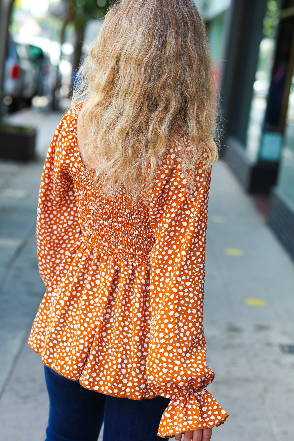 Woman with wavy blonde hair wearing rust smocked top with ruffle hem and white spots walking down city sidewalk