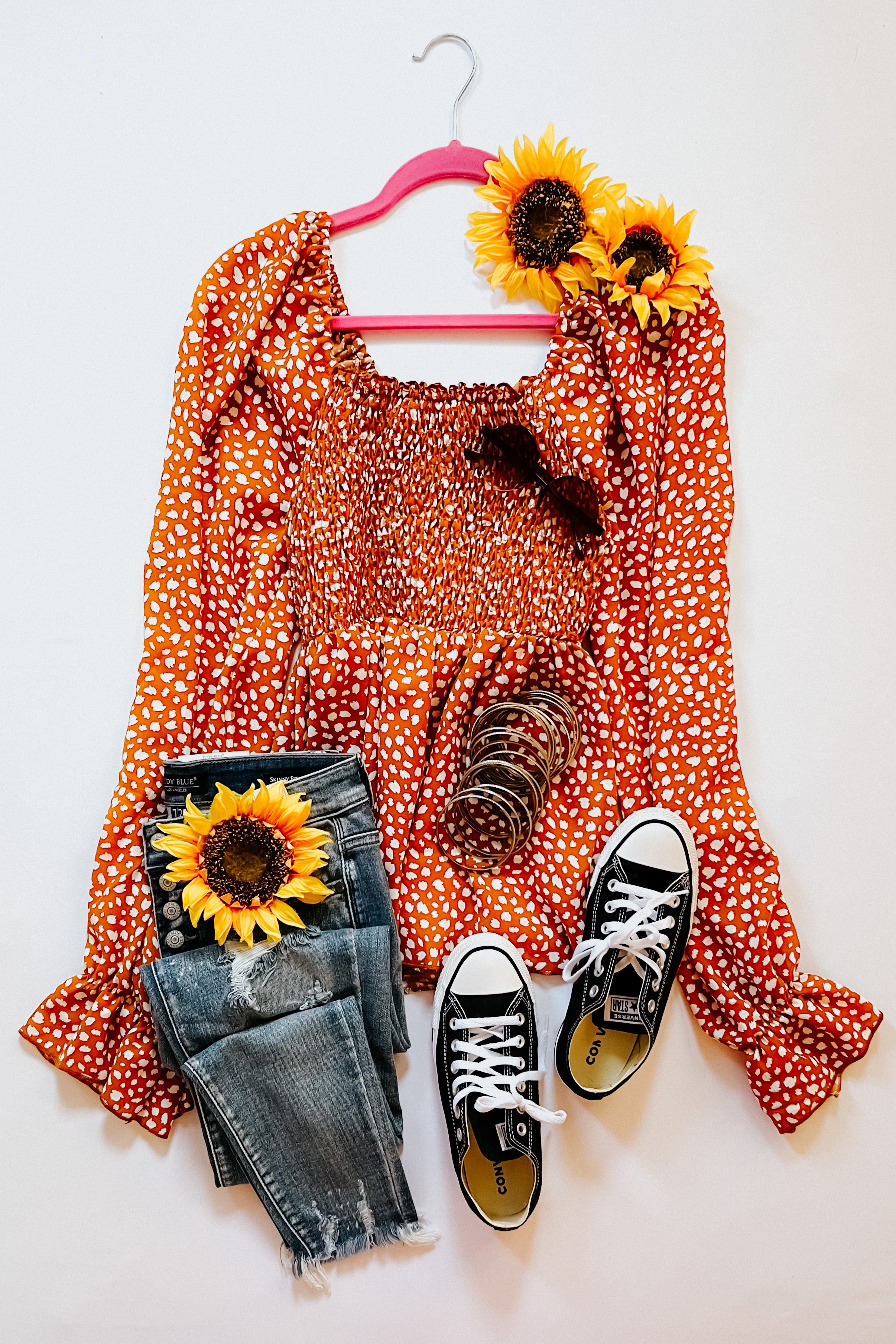 Rust smocked top with ruffle hem styled with distressed jeans, black sneakers, bangles, and sunflowers on white background