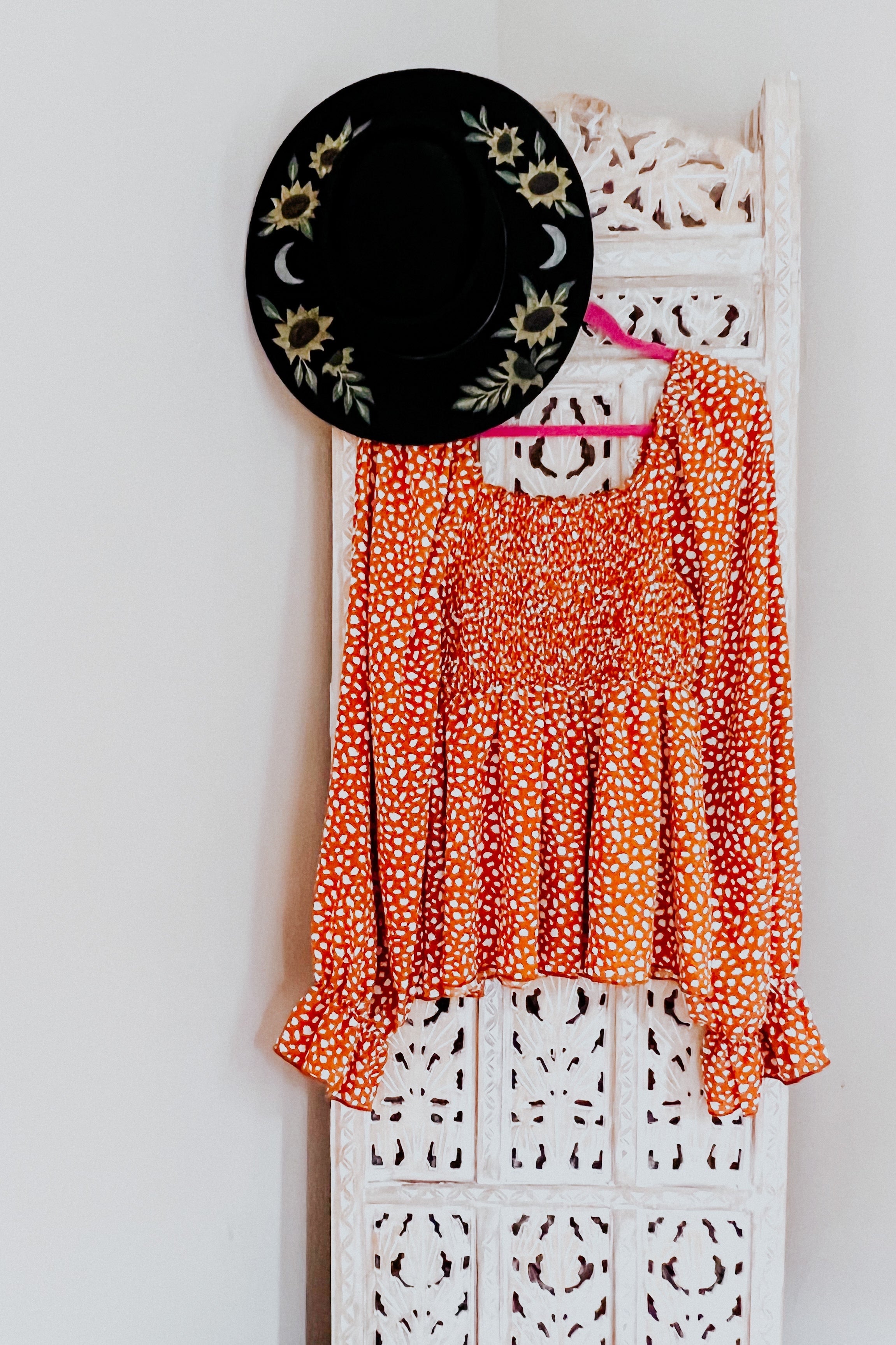 Rust smocked top with ruffle hem hanging on carved white panel beside black hat with sunflower and moon designs