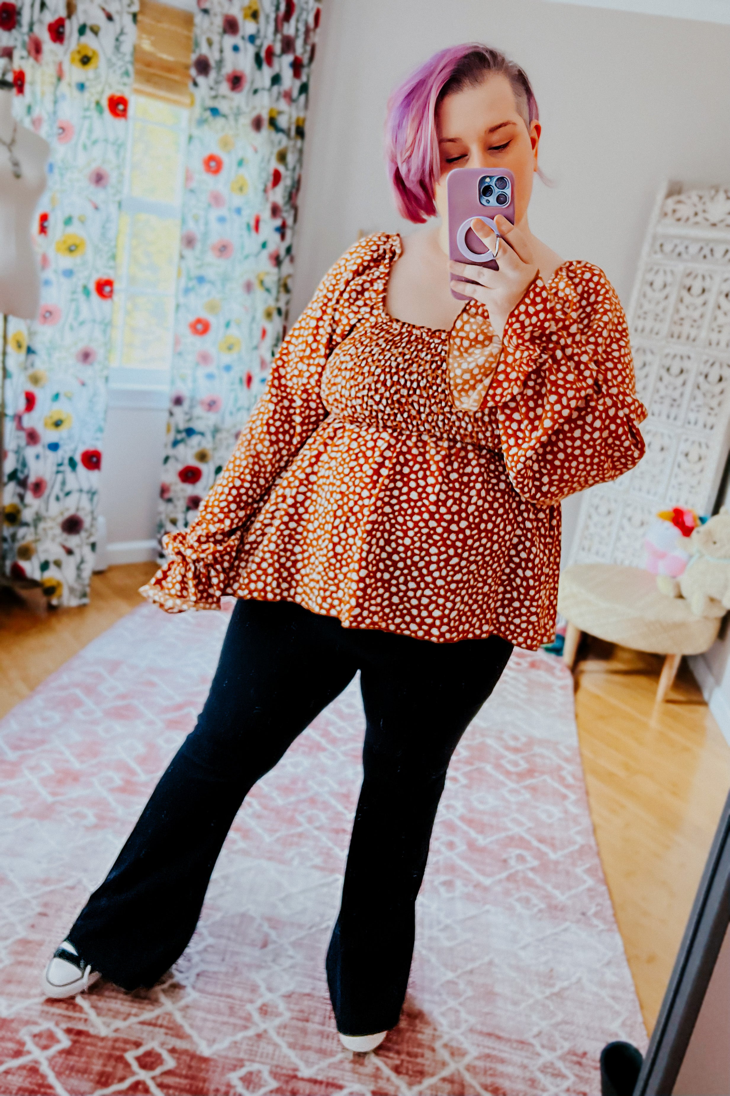 Woman with purple hair wearing rust smocked top with ruffle hem and black flared pants taking mirror selfie indoors