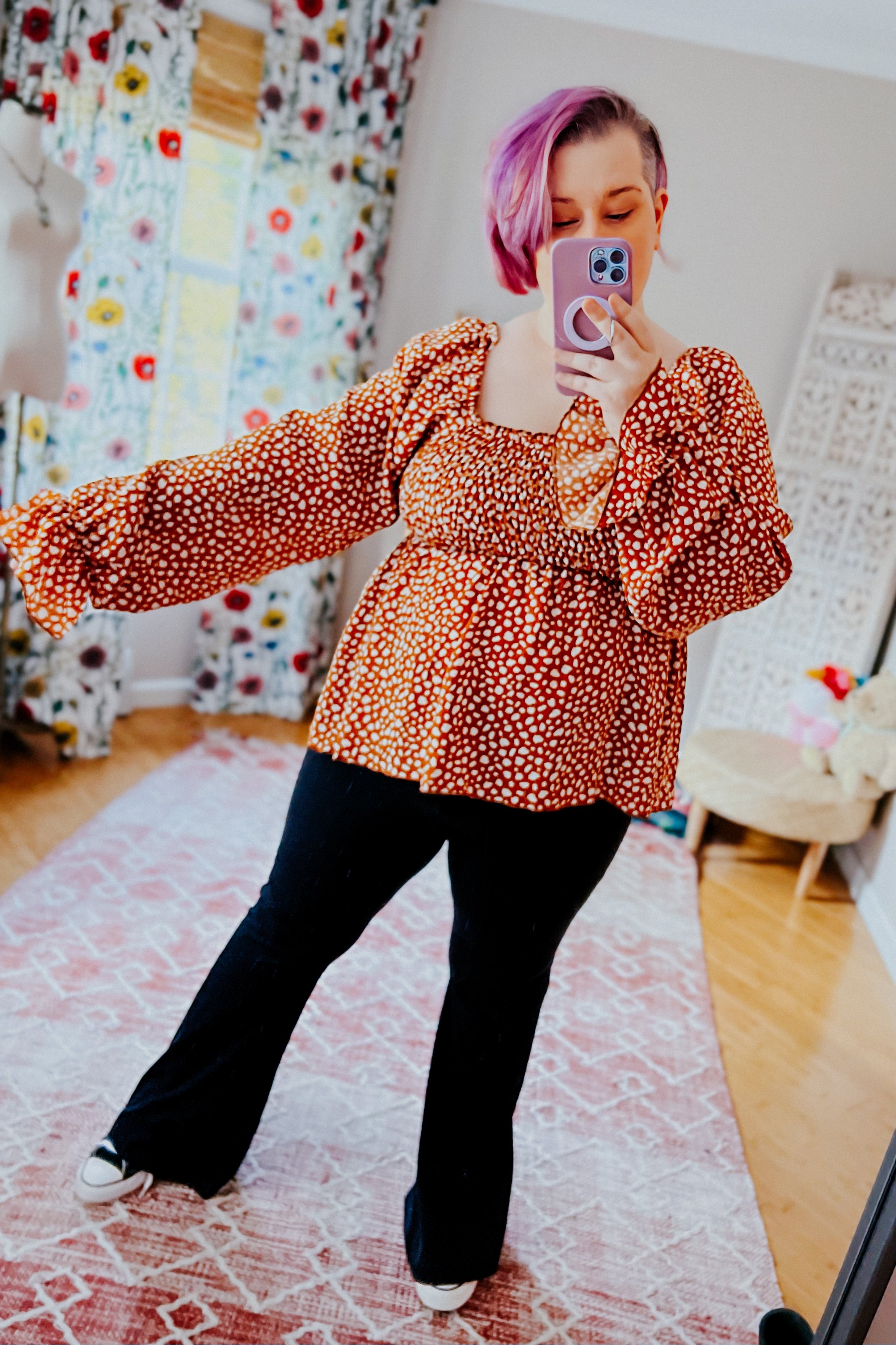 Woman with pink hair wearing rust smocked top with ruffle hem and black pants taking mirror selfie indoors