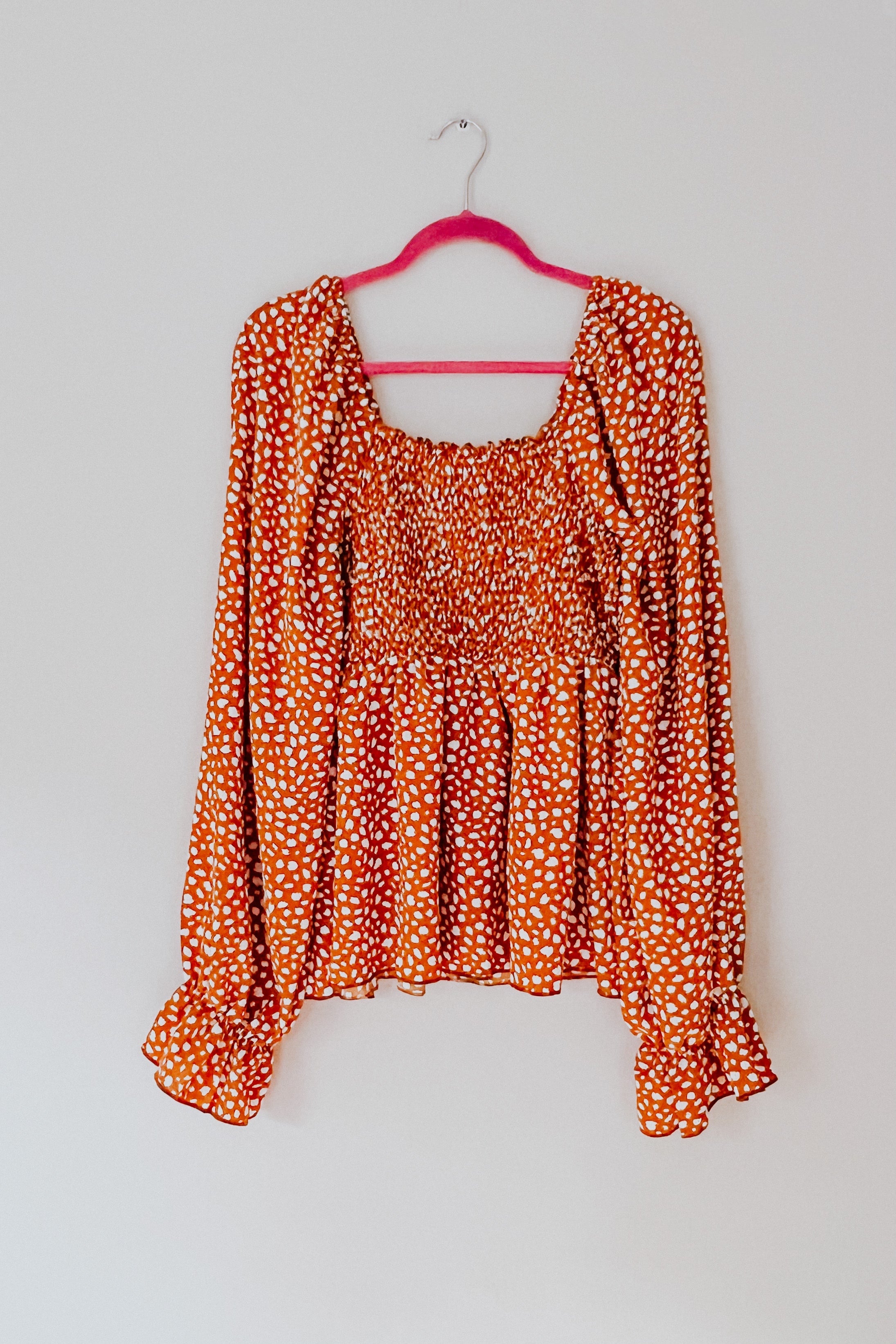 Rust-colored smocked blouse with ruffle hem and long sleeves hanging on pink velvet hanger against white wall