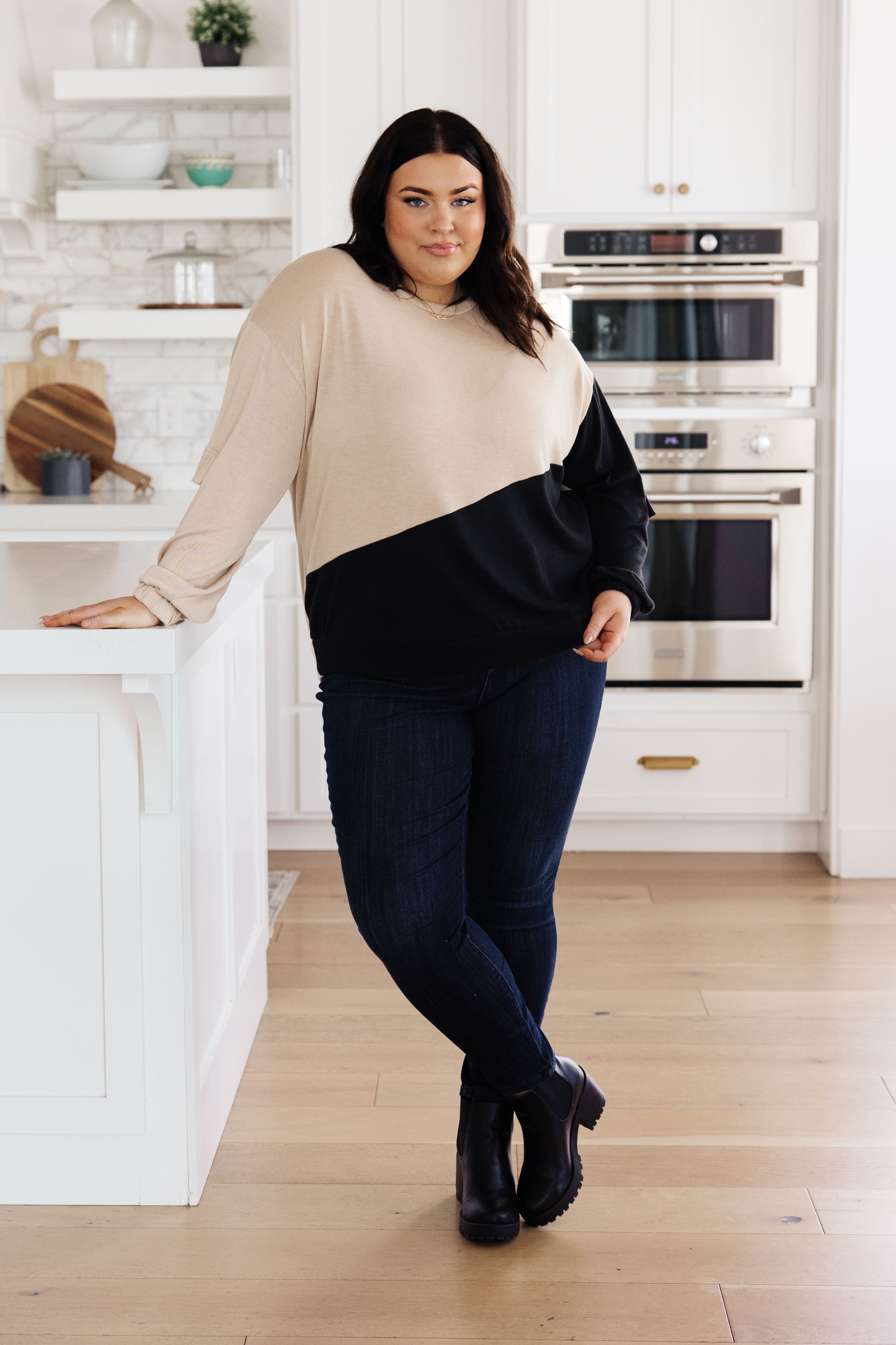 Plus-size model wearing tan and black color block top with cargo sleeves paired with dark jeans and black boots indoors