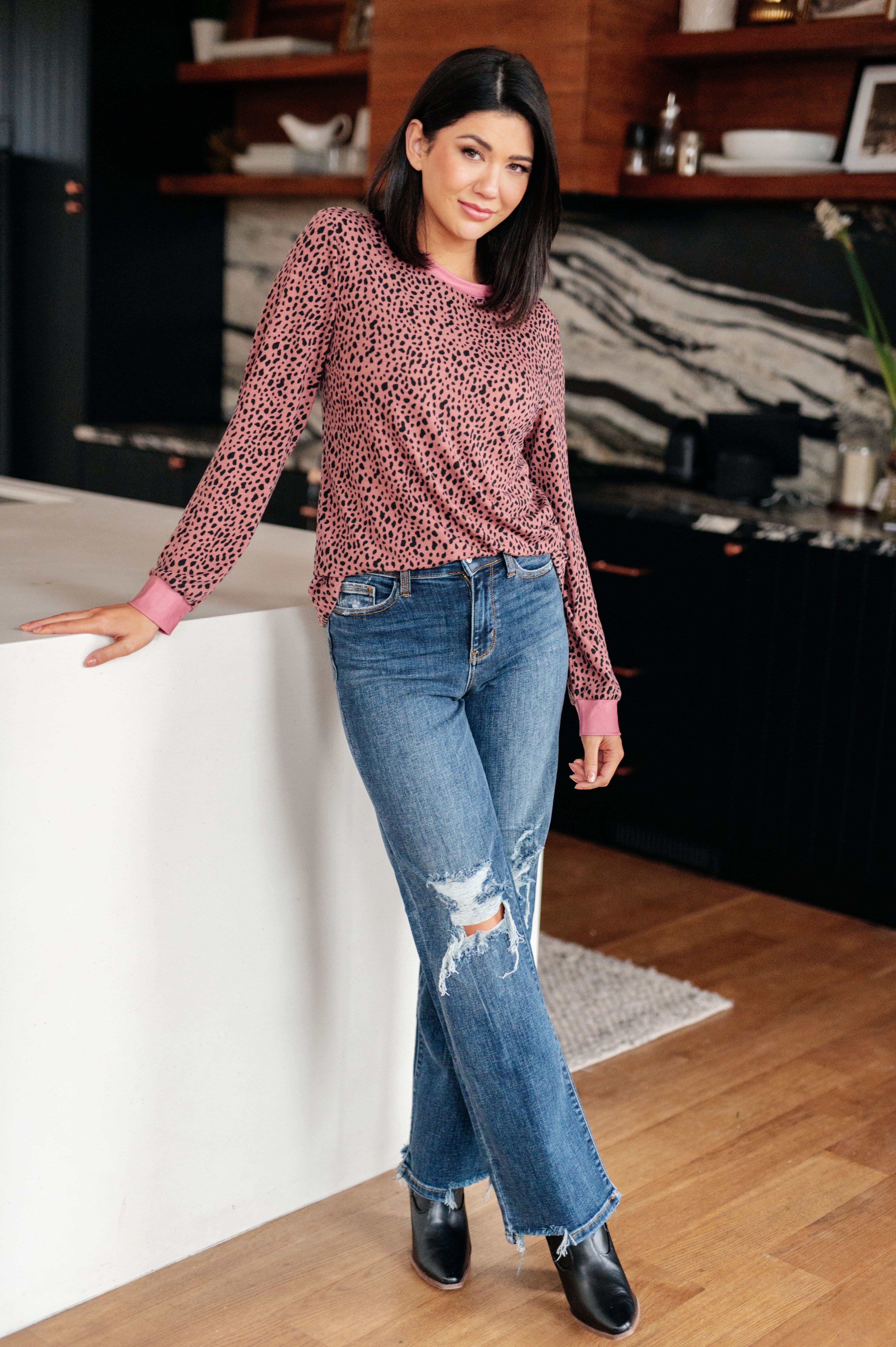 Woman wearing pink spotted long sleeve top styled with distressed blue jeans and black ankle boots in cozy modern kitchen