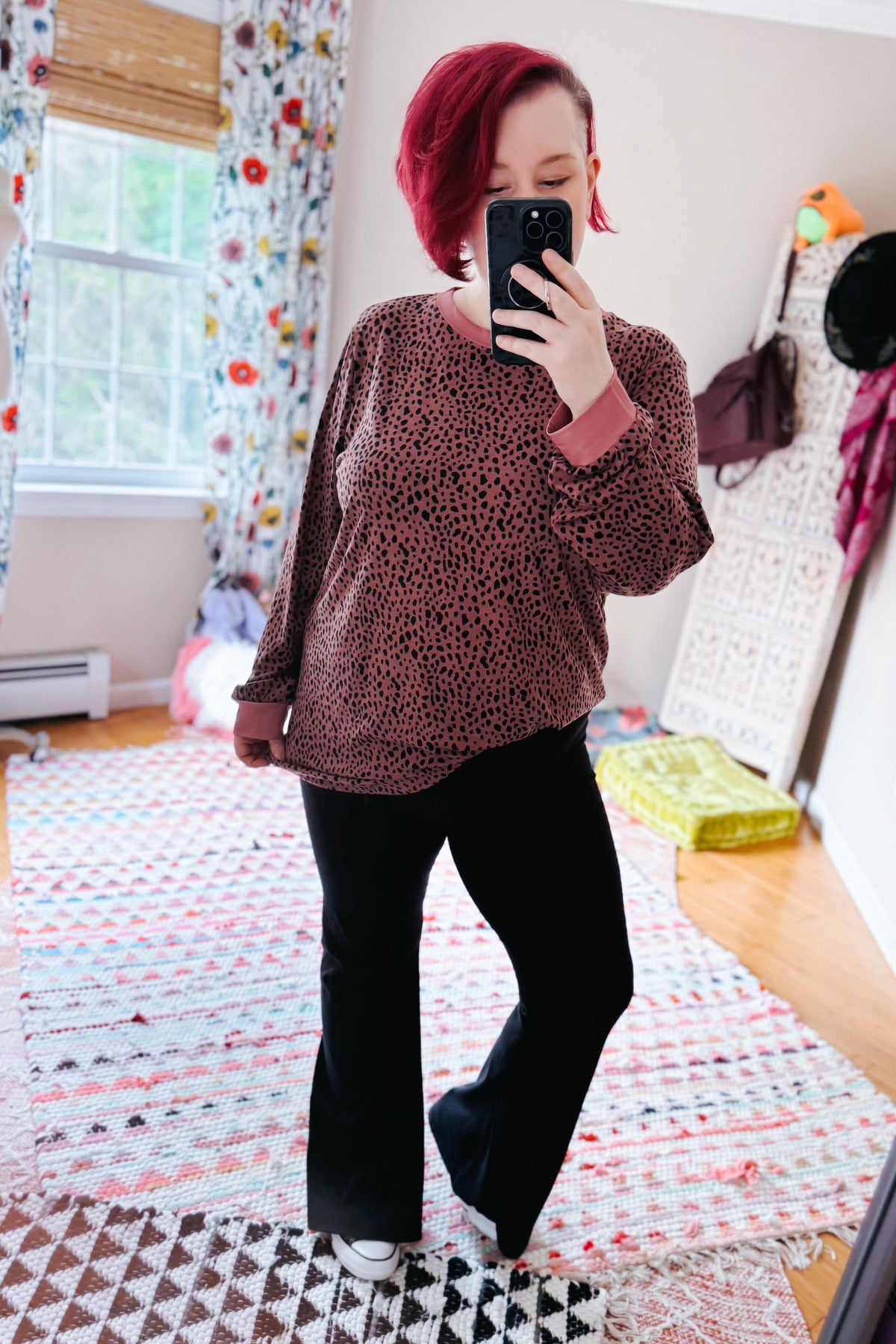 Red haired woman taking a bedroom mirror selfie in a pink spotted long sleeve top black flared pants and white sneakers