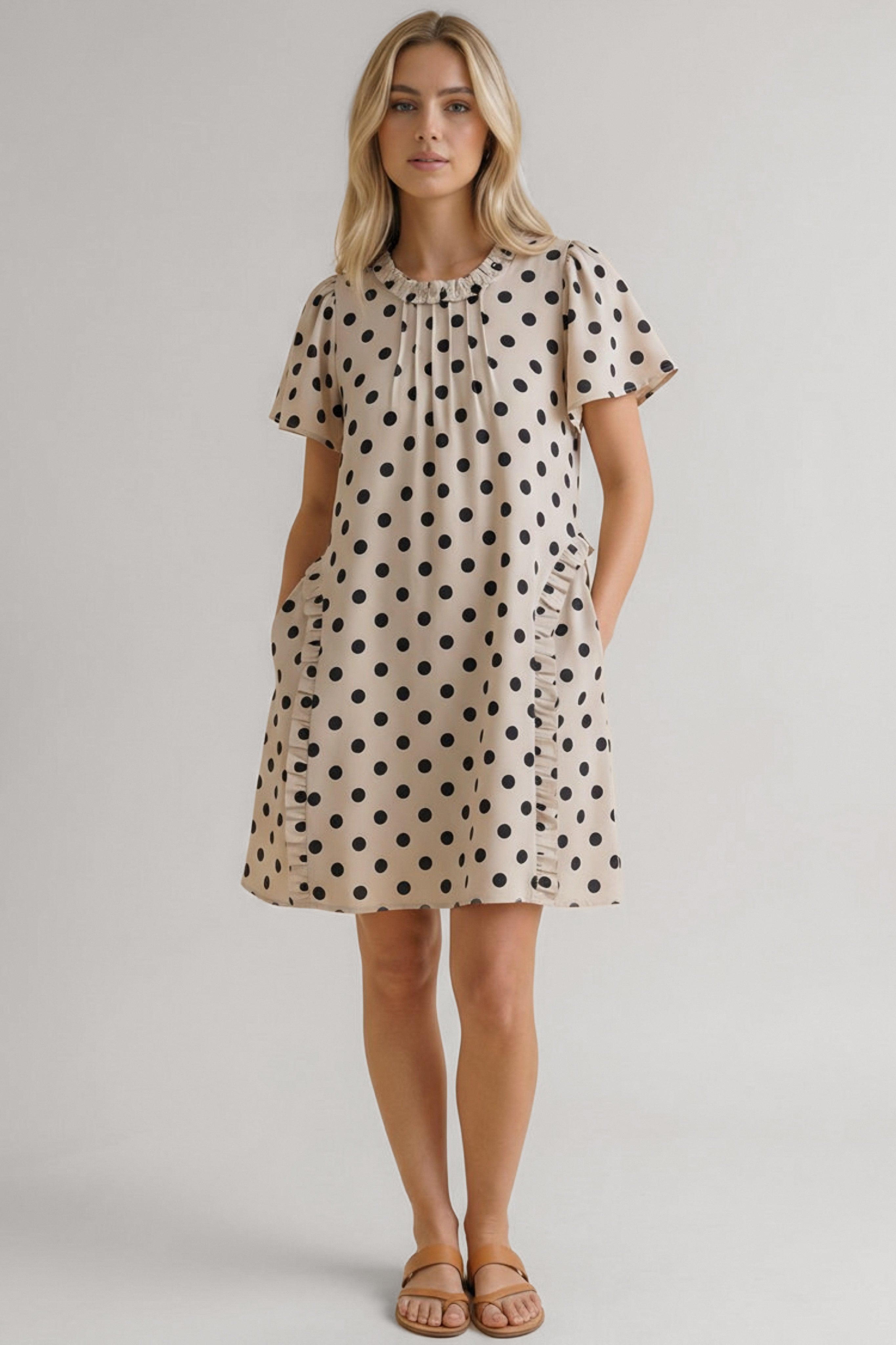 Woman wearing a beige dress with black polka dots, short sleeves, ruffle details near the neckline and pockets, standing facing forward