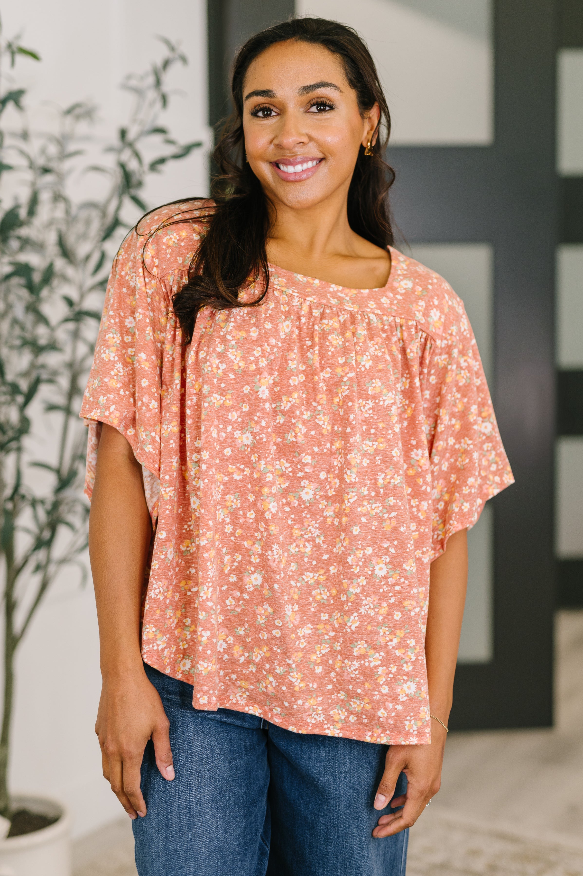 Smiling woman with dark hair wearing a loose-fitting pink floral top and blue jeans standing indoors