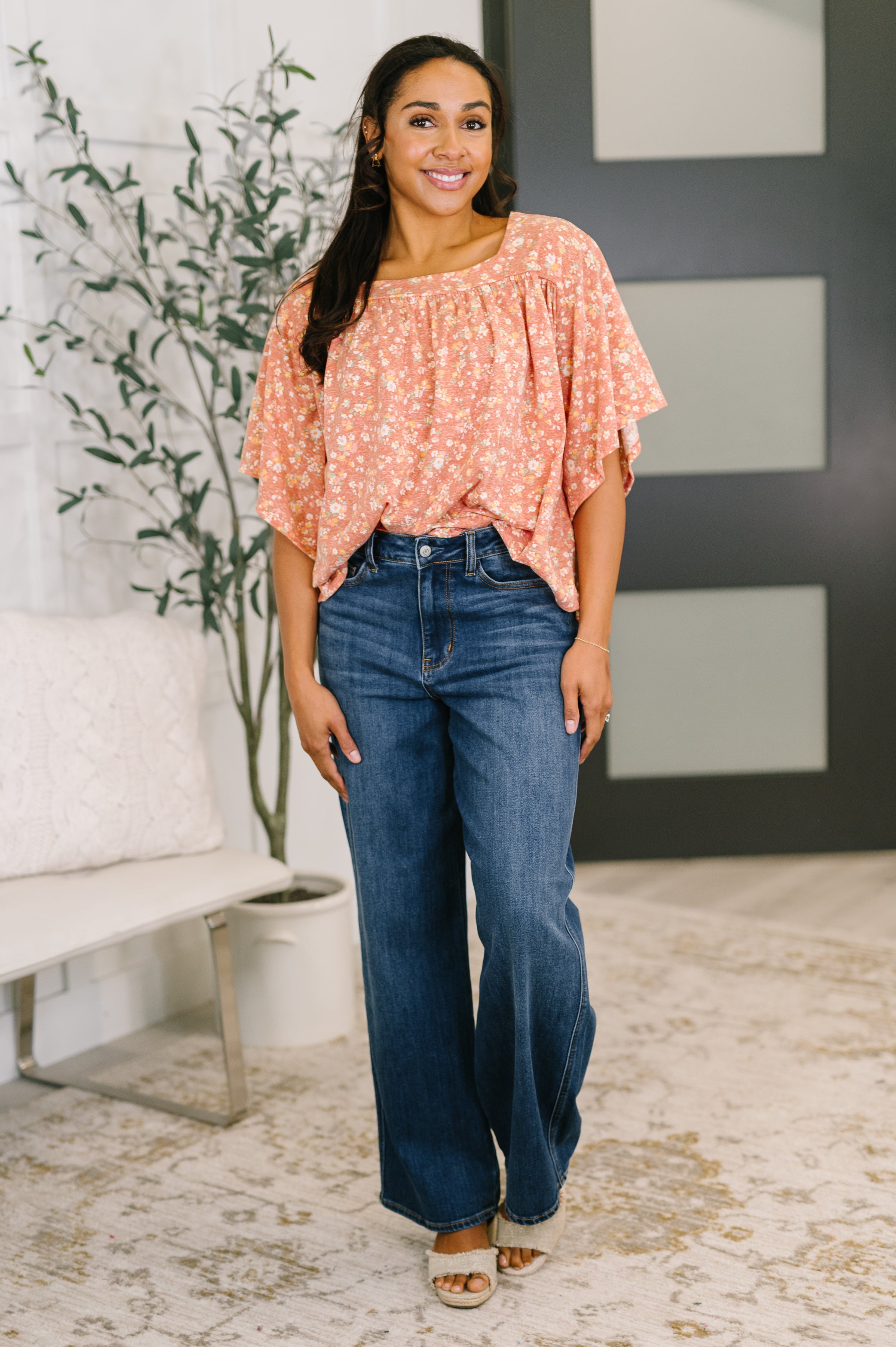 Woman with long dark hair wearing a loose-fitting pink floral top with wide sleeves and high-waisted blue jeans