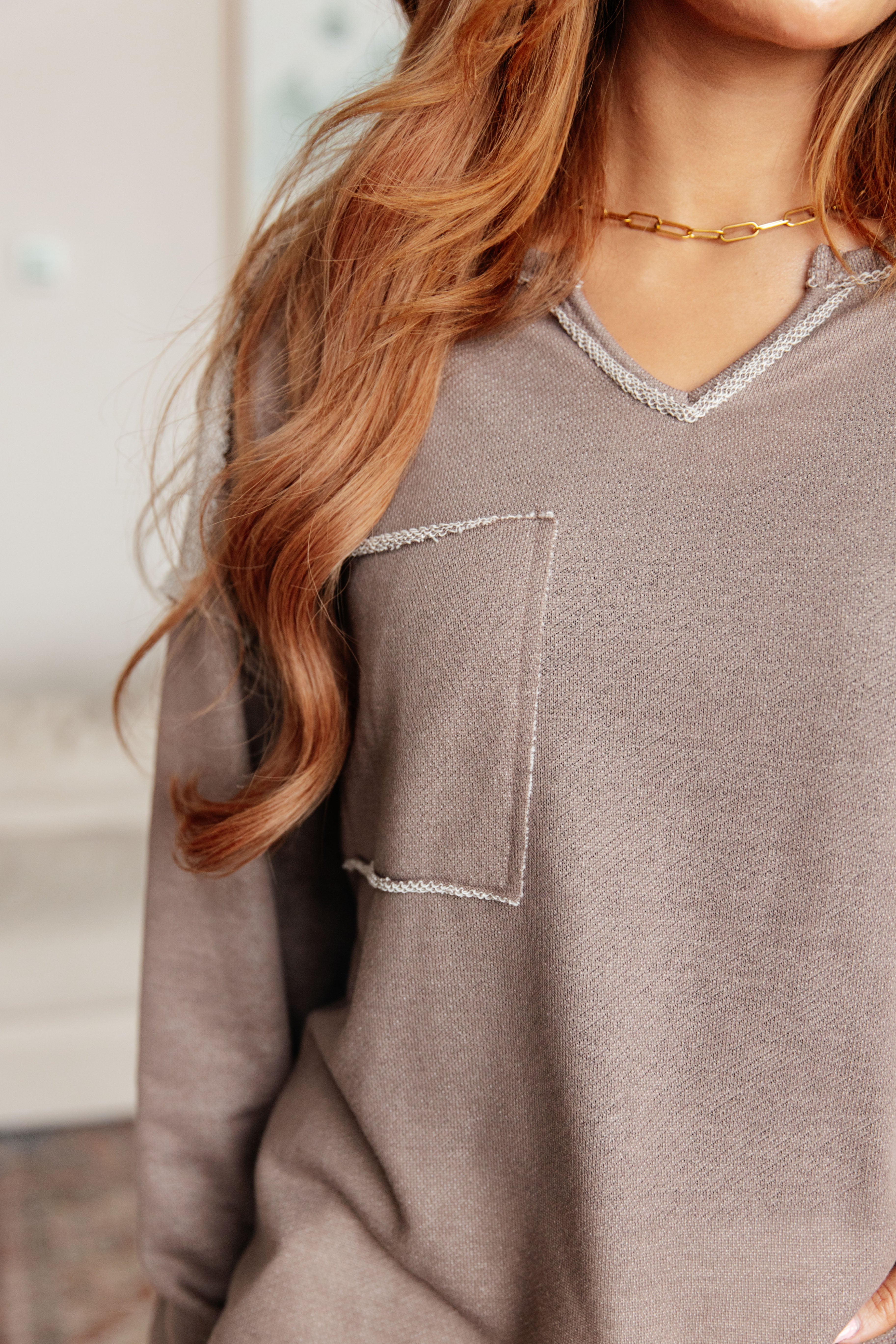 Woman wearing a taupe reverse stitch v-neck pullover with raw accents standing in an indoor setting.