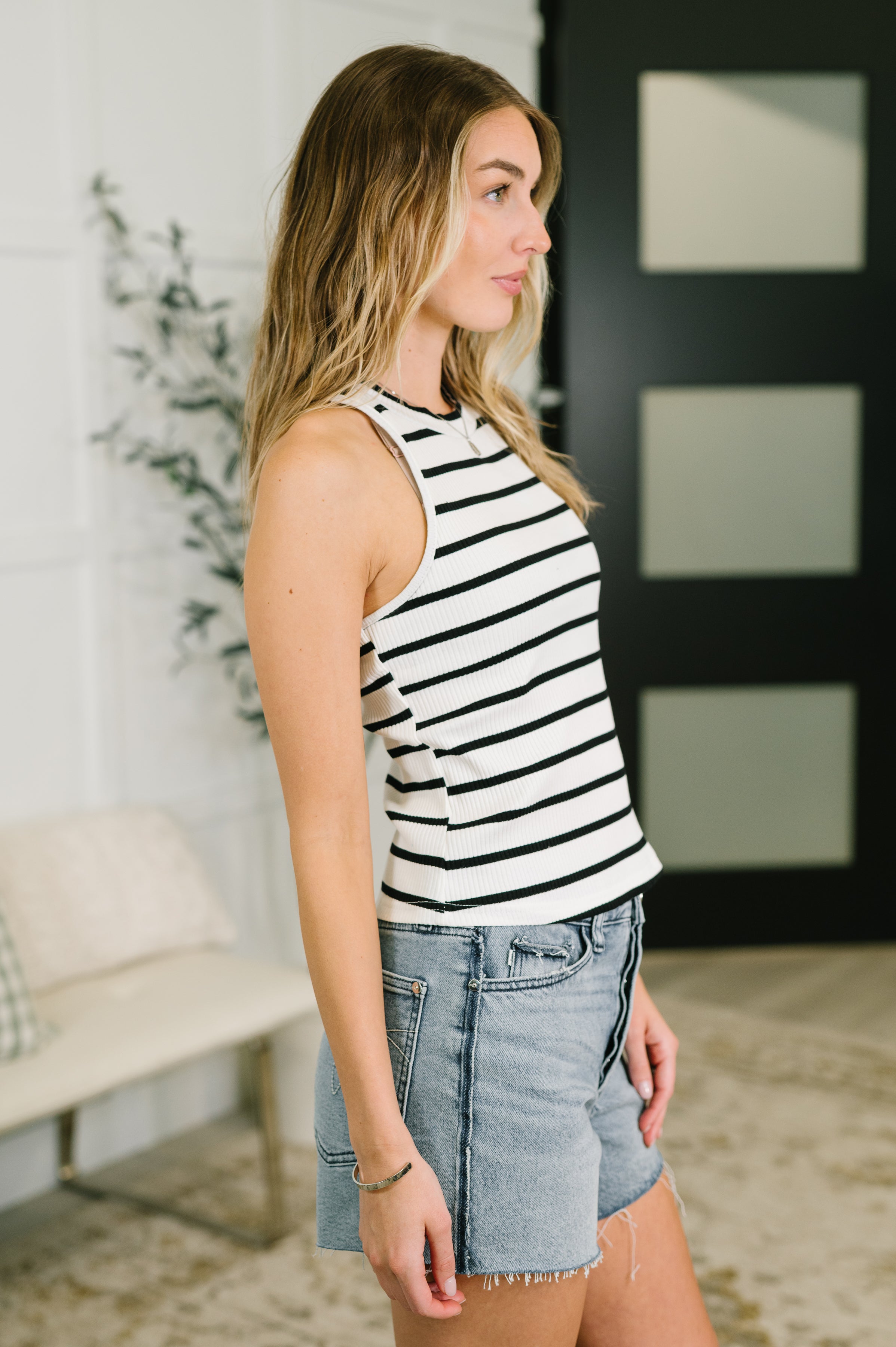 Side profile of woman wearing ribbed ivory halter tank with thin black stripes and light denim shorts indoors