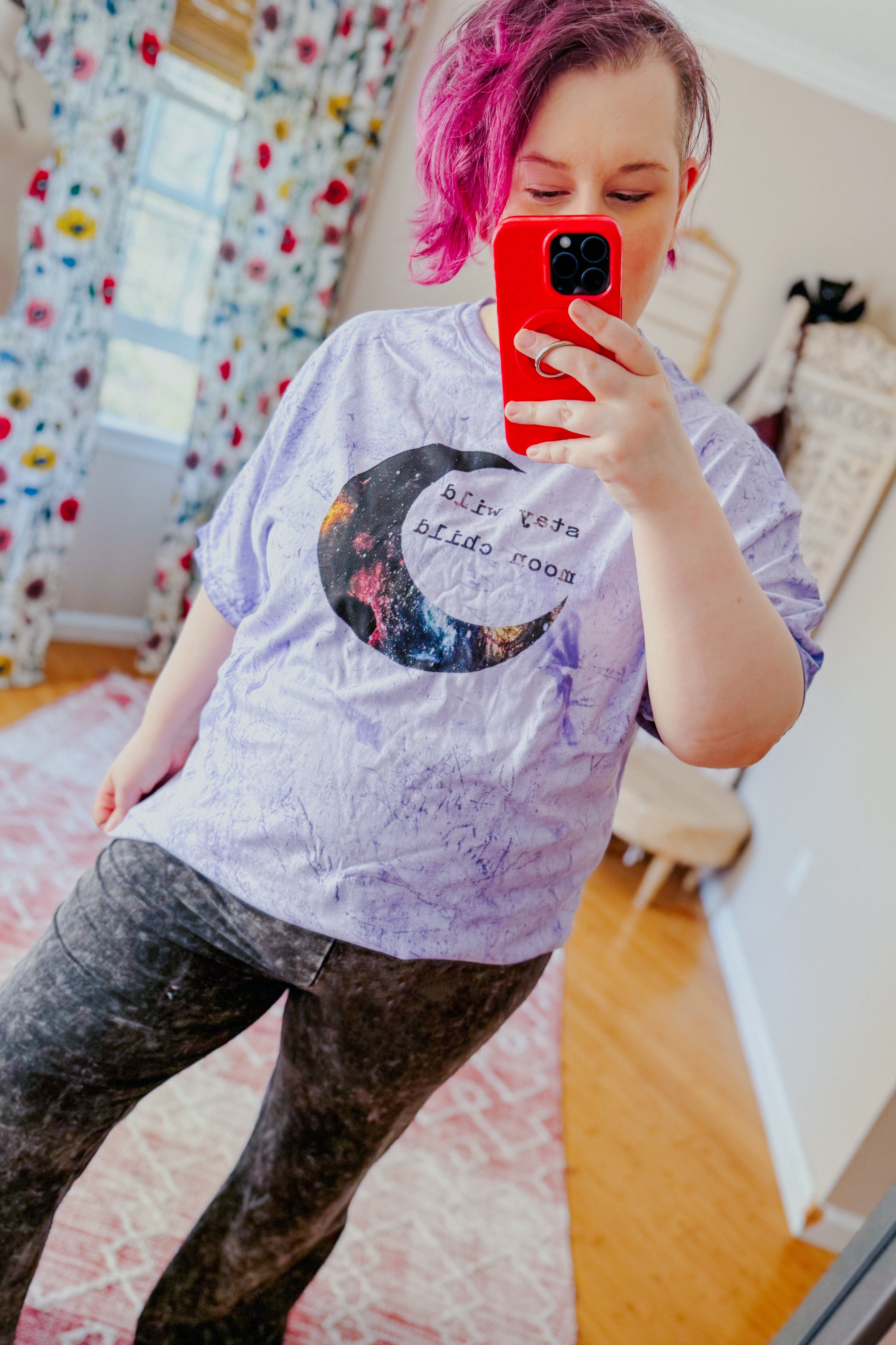 Woman with short magenta hair wearing a purple tie-dye tee with a crescent moon graphic and black leggings indoors